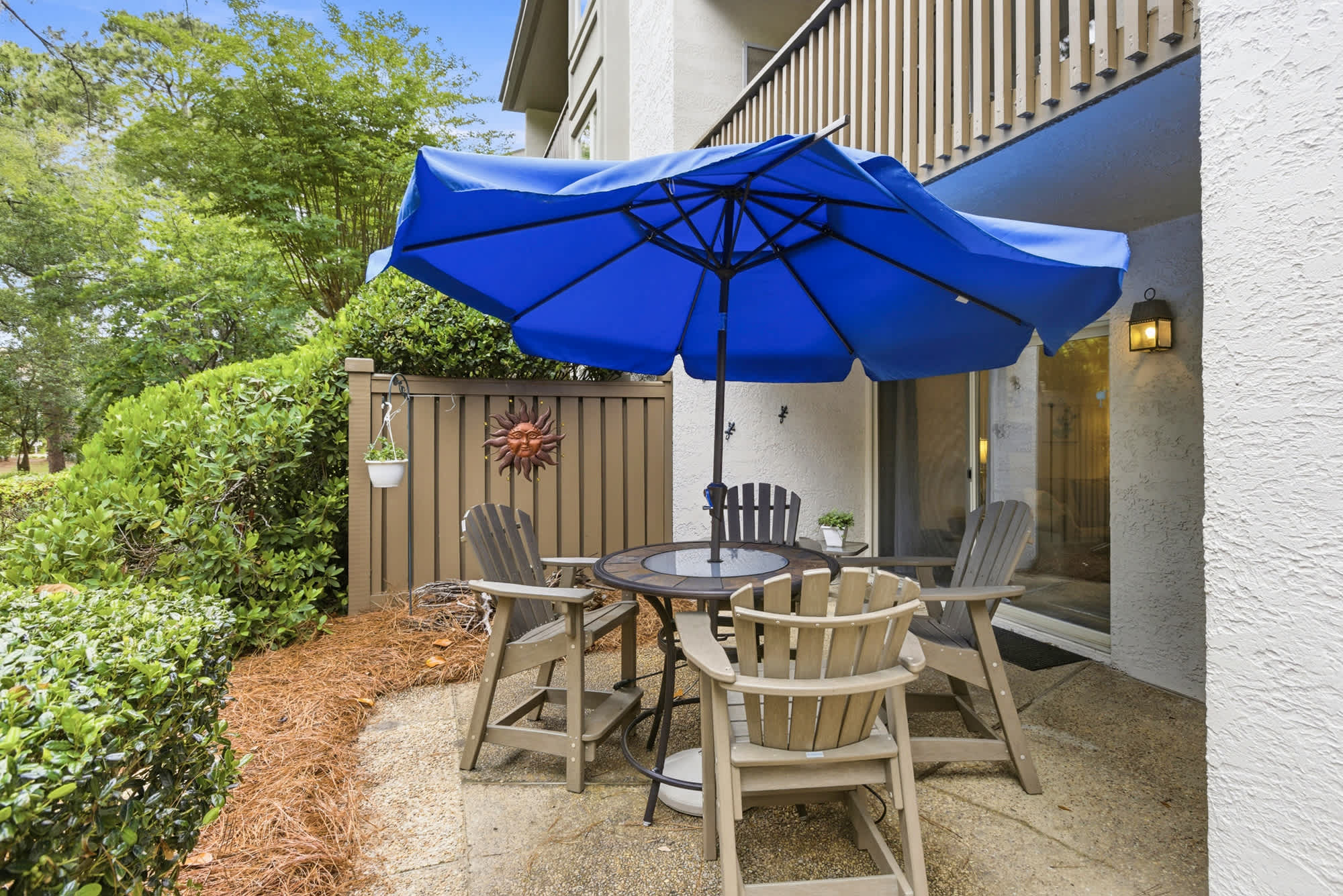 Vivid sapphire shades from the broad umbrella cast a refreshing glow over this stone patio. Weathered adirondack chairs gather around an intimate table, creating a peaceful corner for slow mornings and quiet conversation among the greenery.