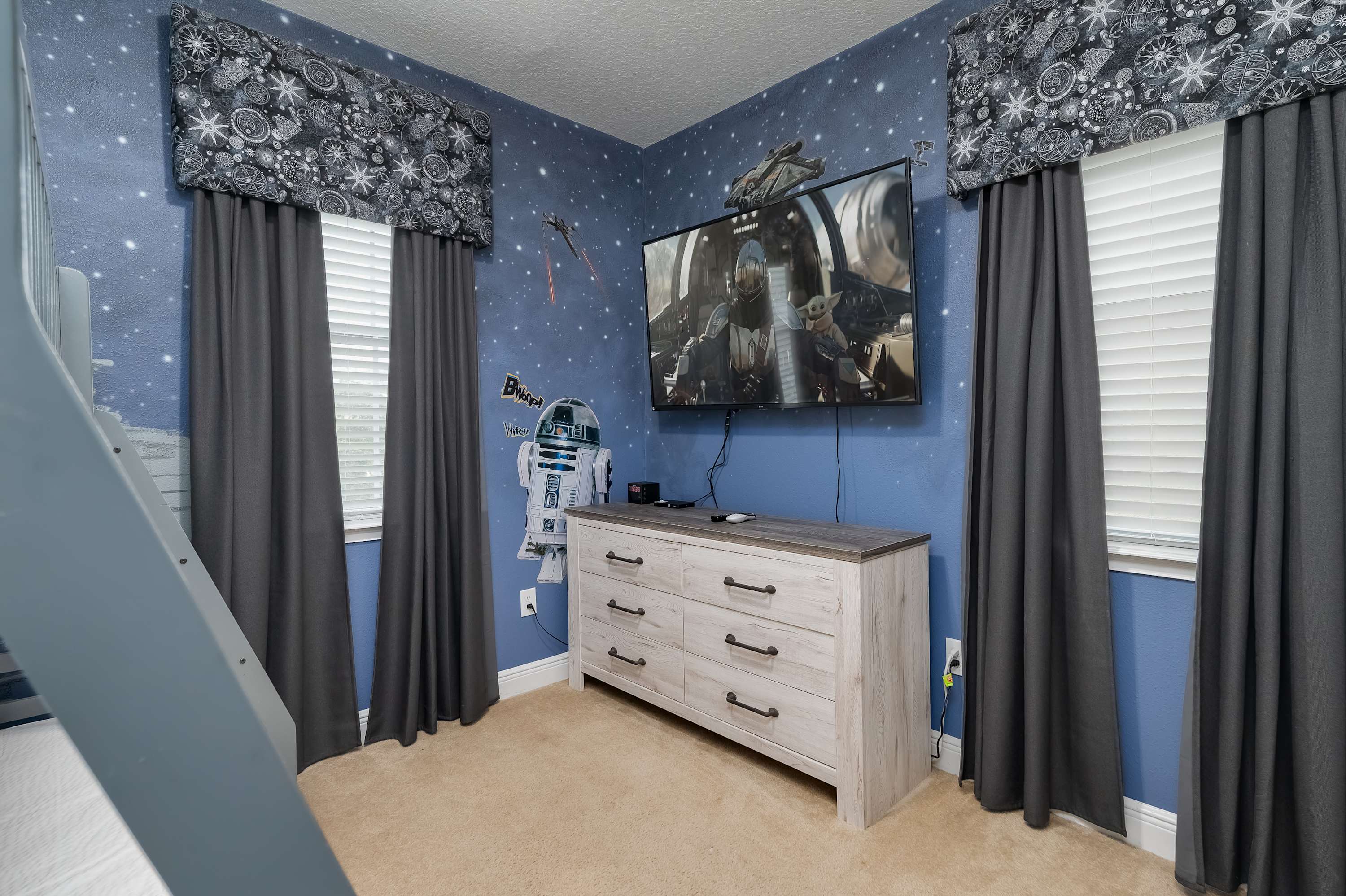 Star wars-themed room with a wall-mounted television and dresser between two windows with dark curtains.