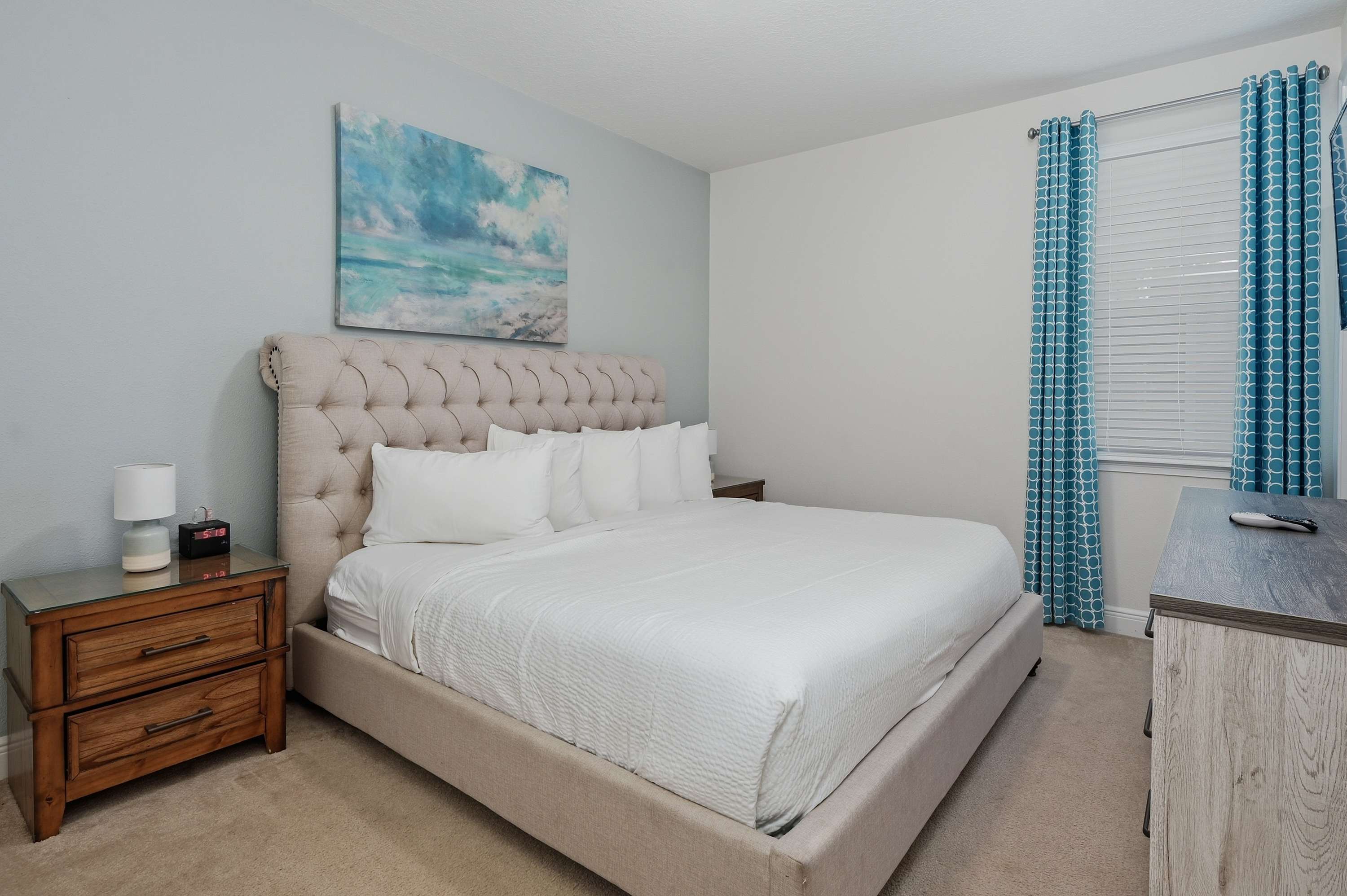 Bedroom with a tufted bedframe, ocean-themed artwork, and patterned blue curtains.