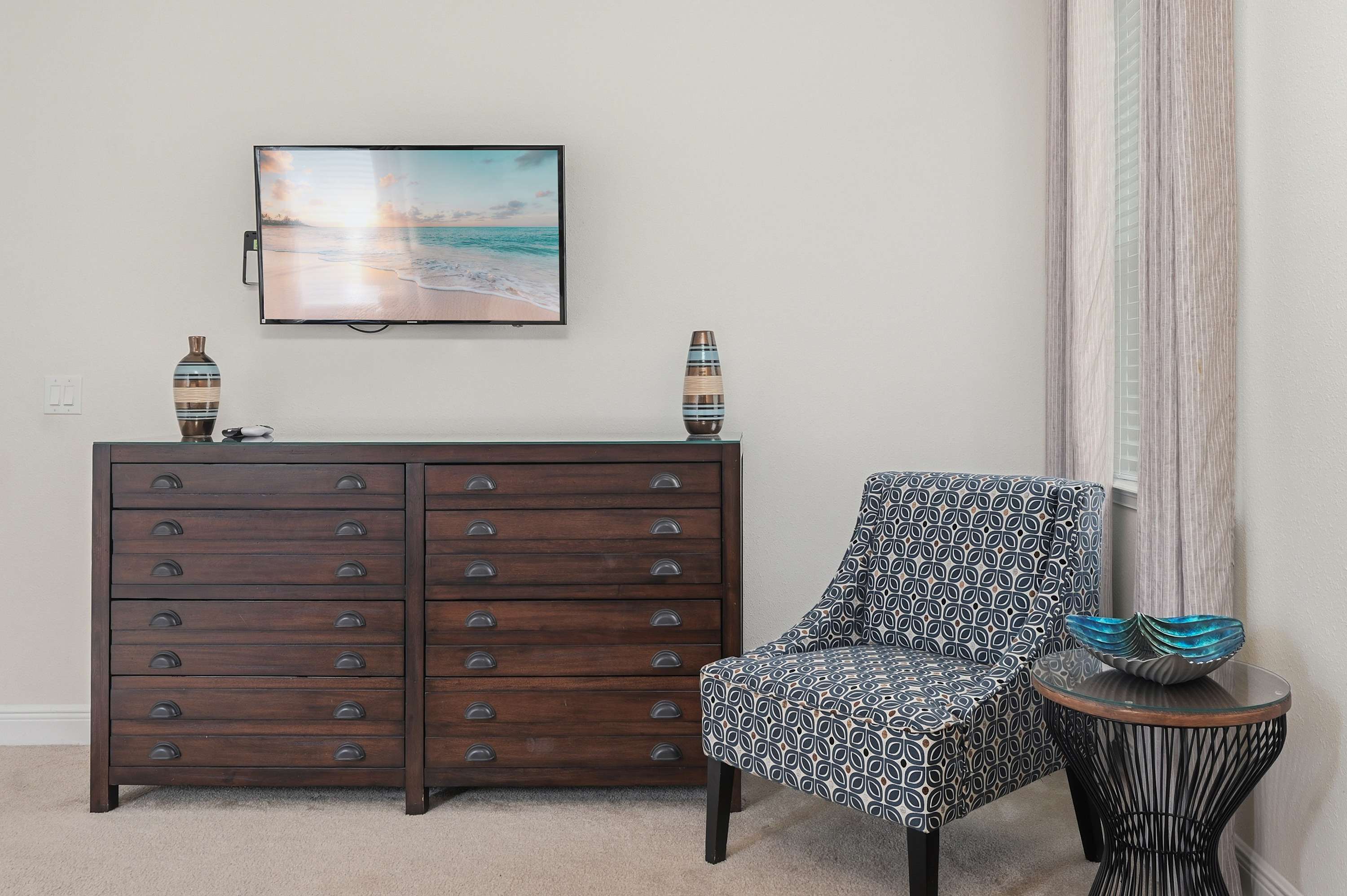 A patterned armchair and decorative table complement the dark wood dresser with a mounted flat-screen television.