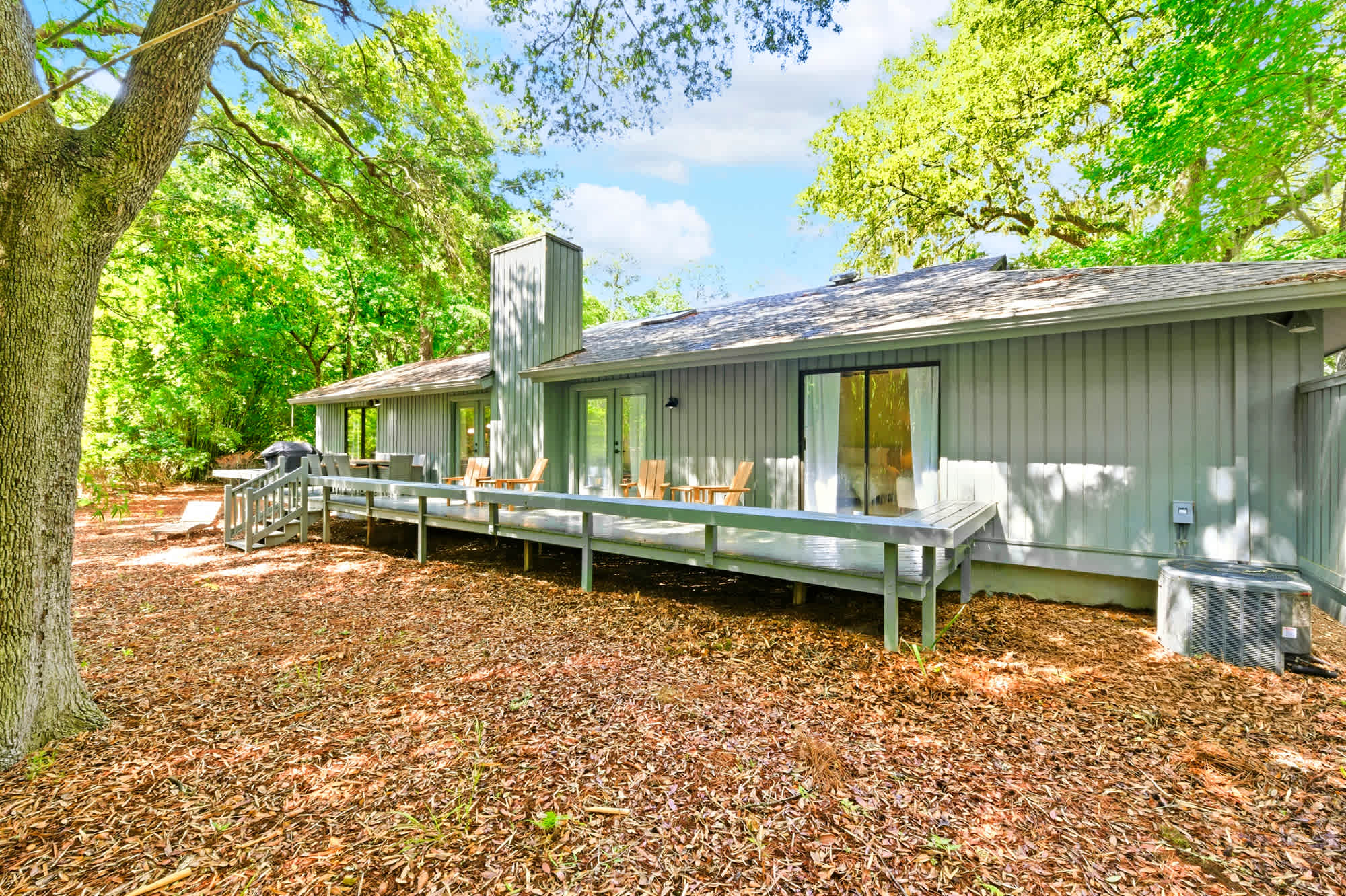 The home's private deck stretches beneath a canopy of ancient oaks, offering a serene spot for morning coffee. This secluded porch blends soft grey siding with the vibrant greens of the forest to create a quiet, airy escape.