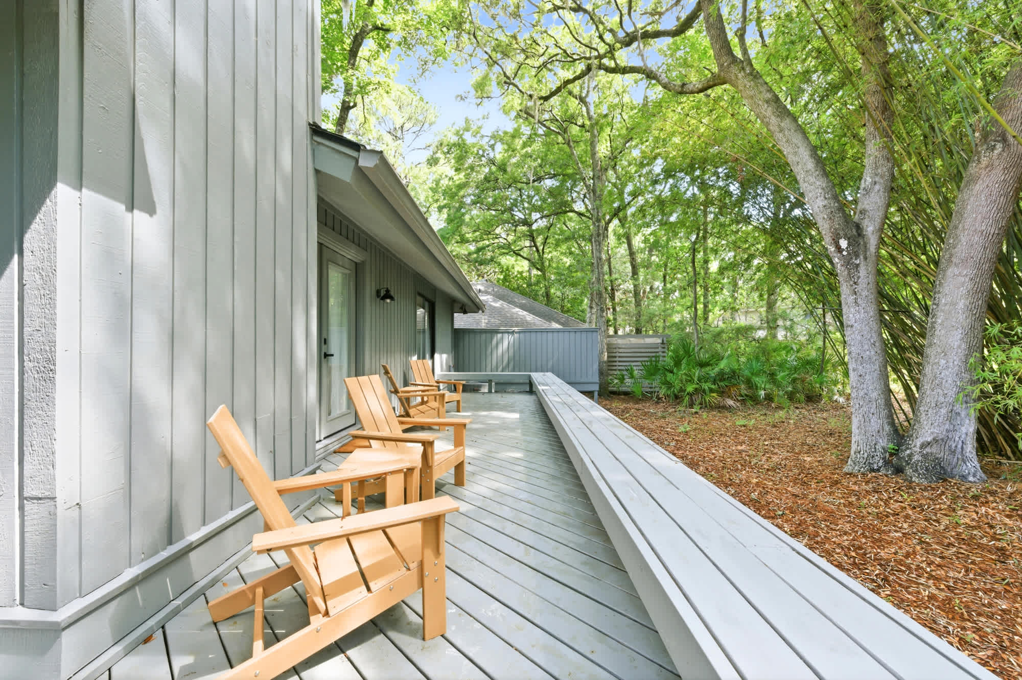 Golden wooden chairs line this expansive gray deck, offering a quiet front row seat to the lush canopy above. Dappled afternoon brightness filters through the towering oaks, creating a serene space for coffee amidst the trees.