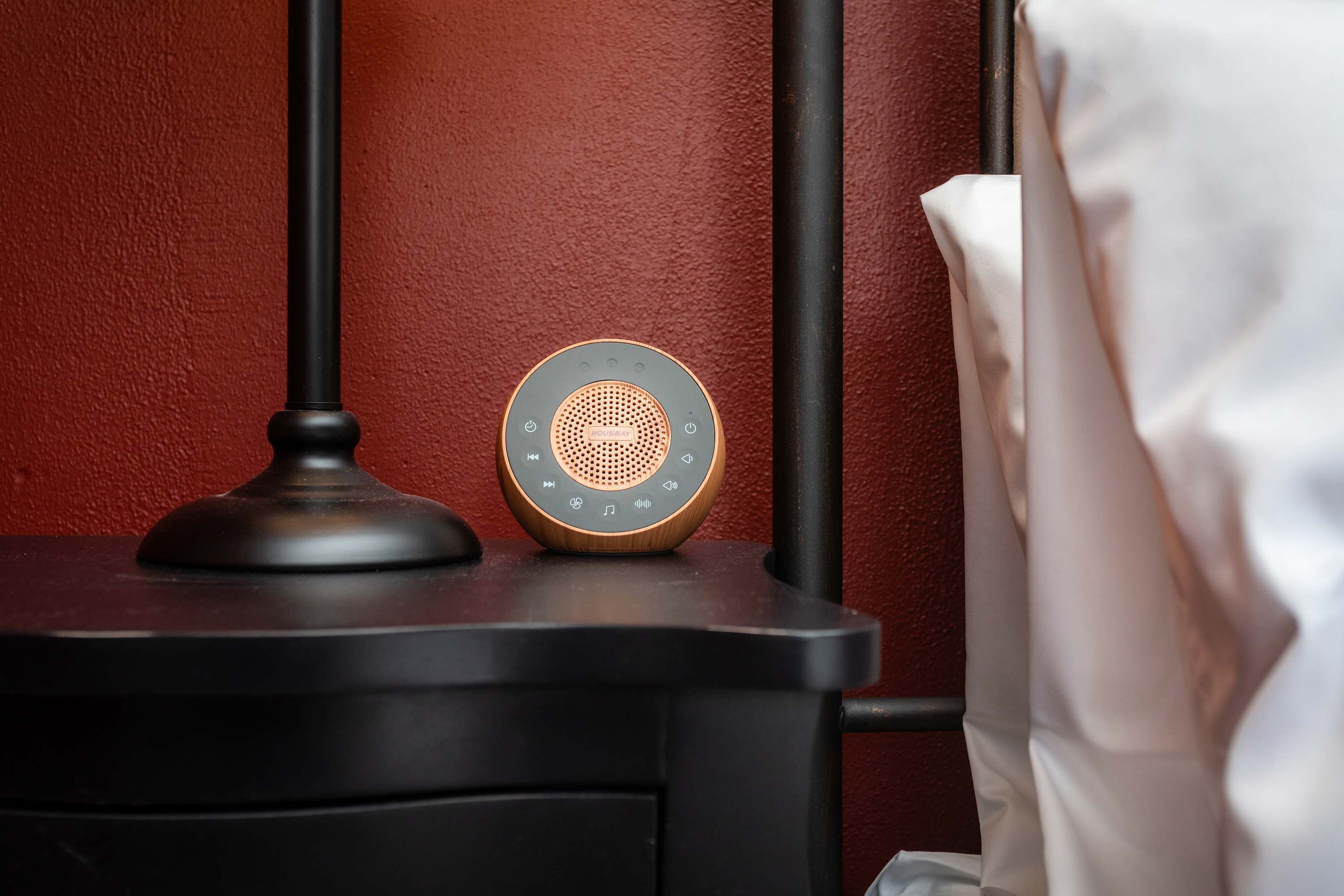 Compact speaker on a bedside table with a black lamp and red textured wall for a cozy atmosphere.