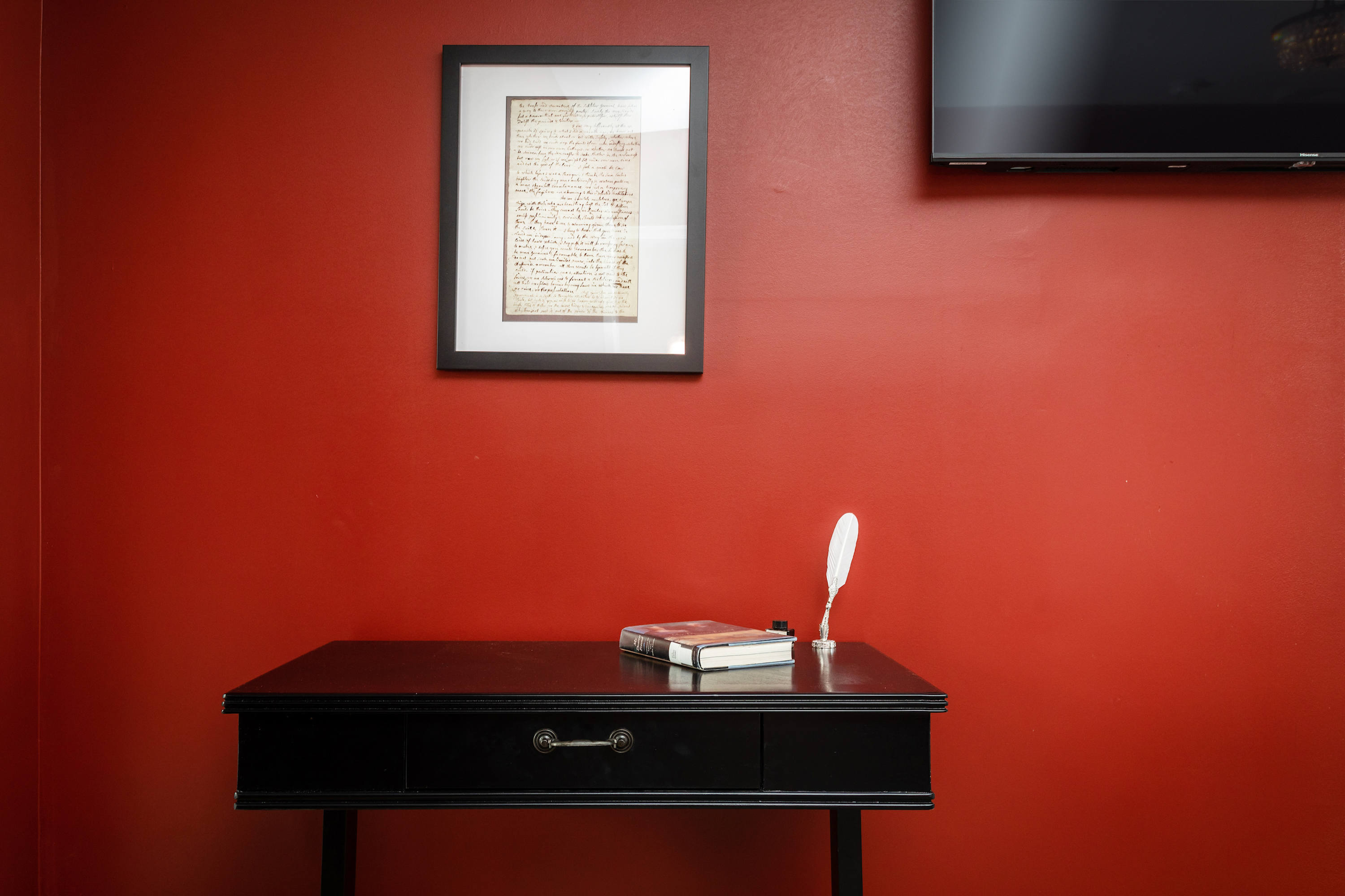 A black desk with a book and quill set against a red wall with framed artwork and a mounted television.