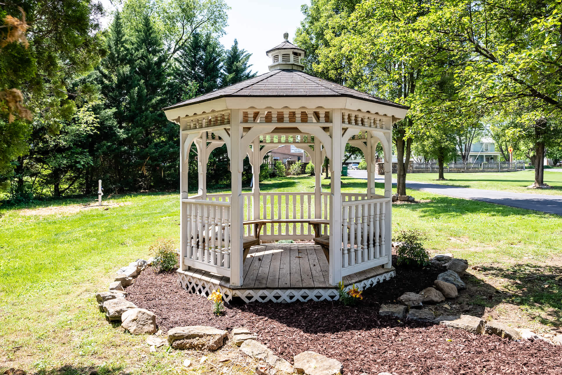 A white wooden gazebo surrounded by lush greenery and a stone-bordered flower bed.