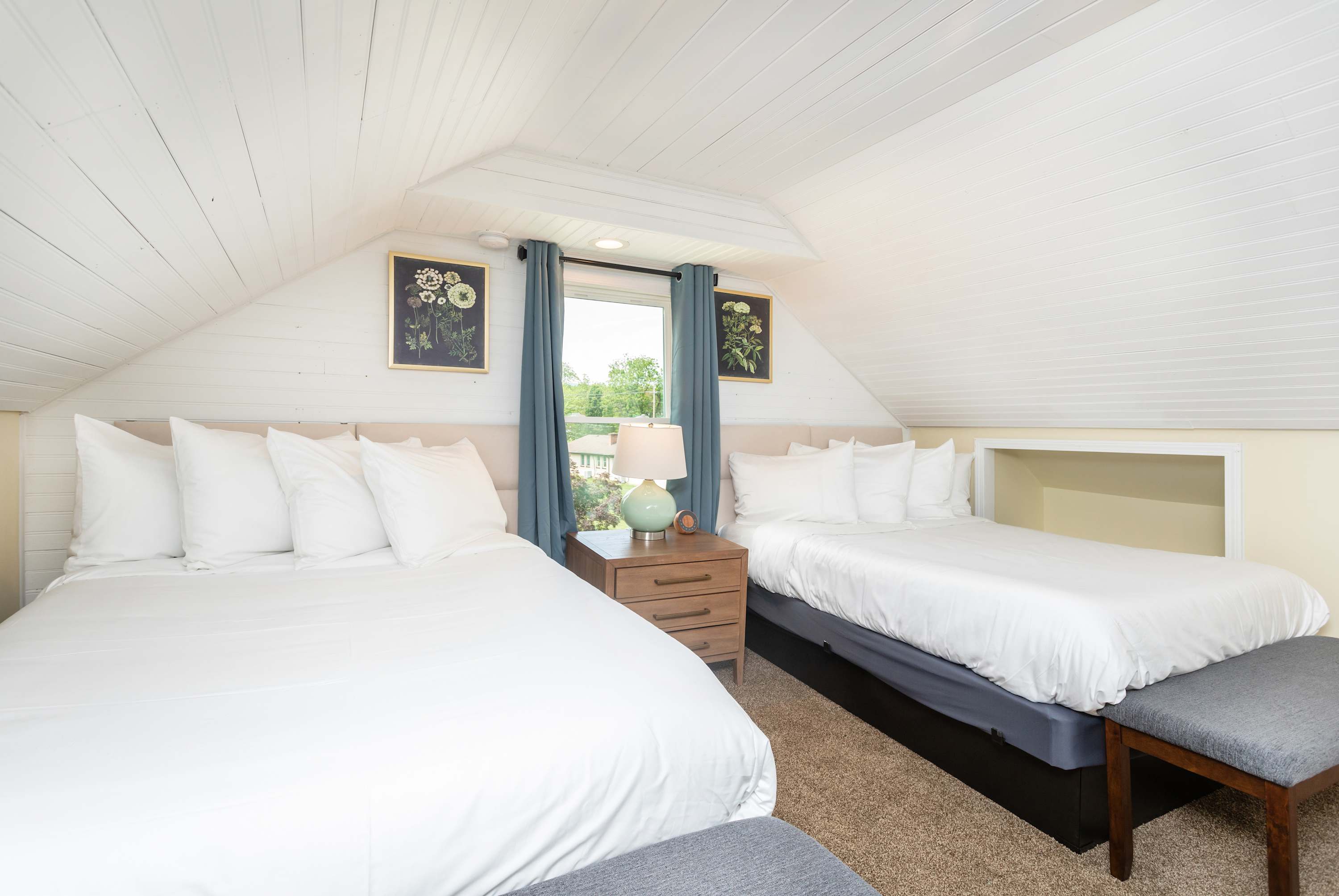 Two queen beds with white linens and a window with blue curtains in a cozy attic bedroom