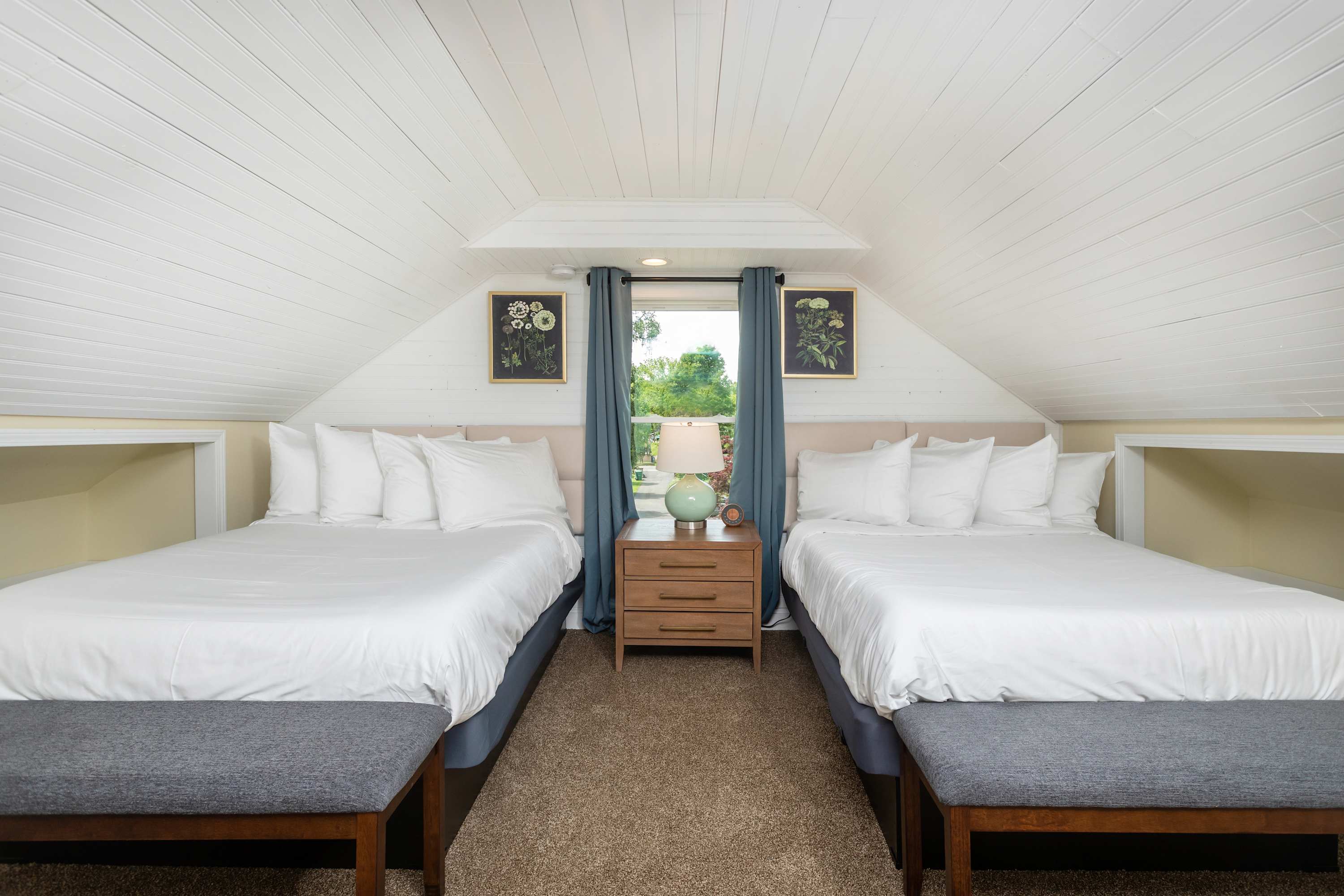 Two queen beds with a nightstand and lamp in a bright attic bedroom.
