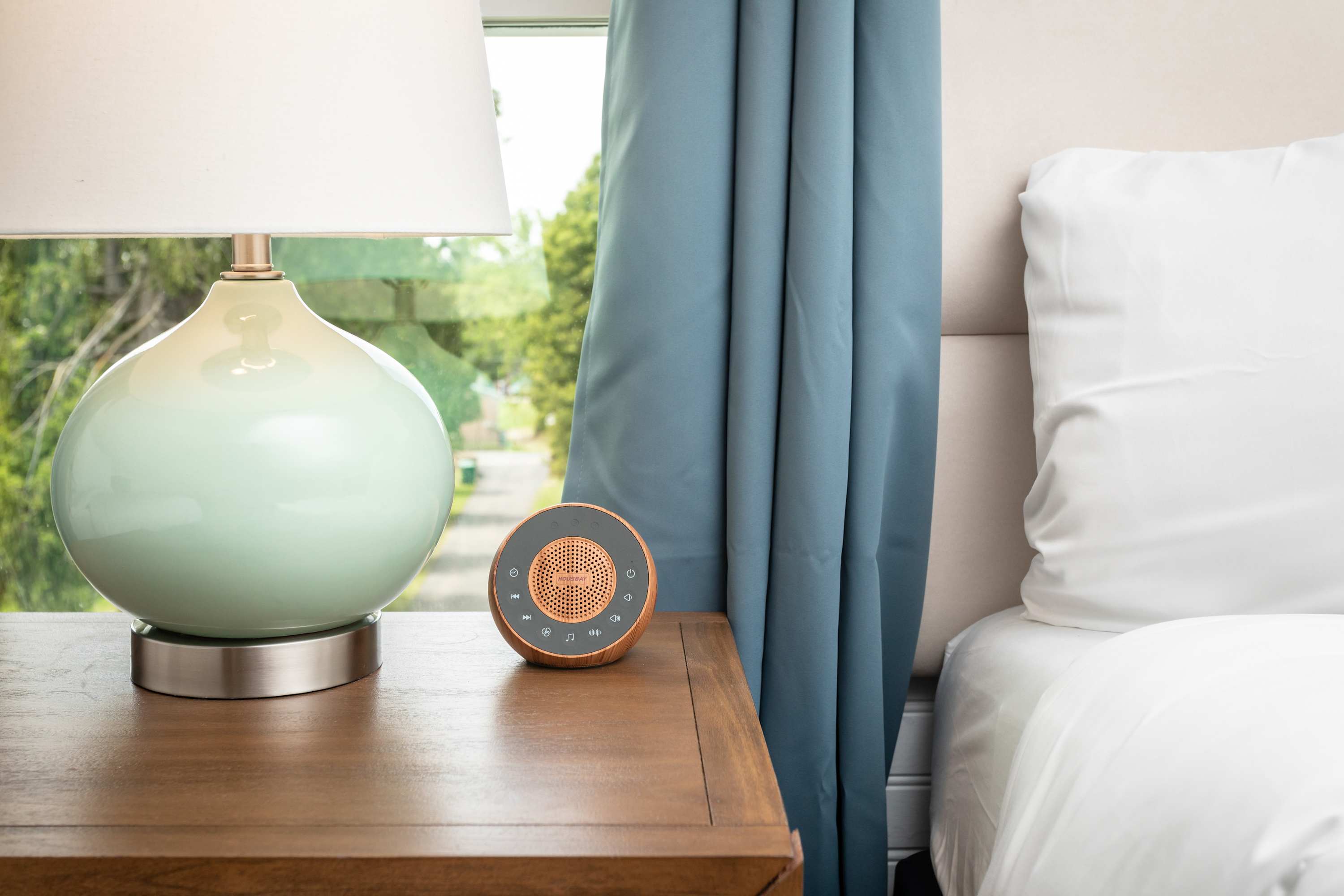 A bedside table with a modern lamp and a compact speaker next to a neatly made bed.