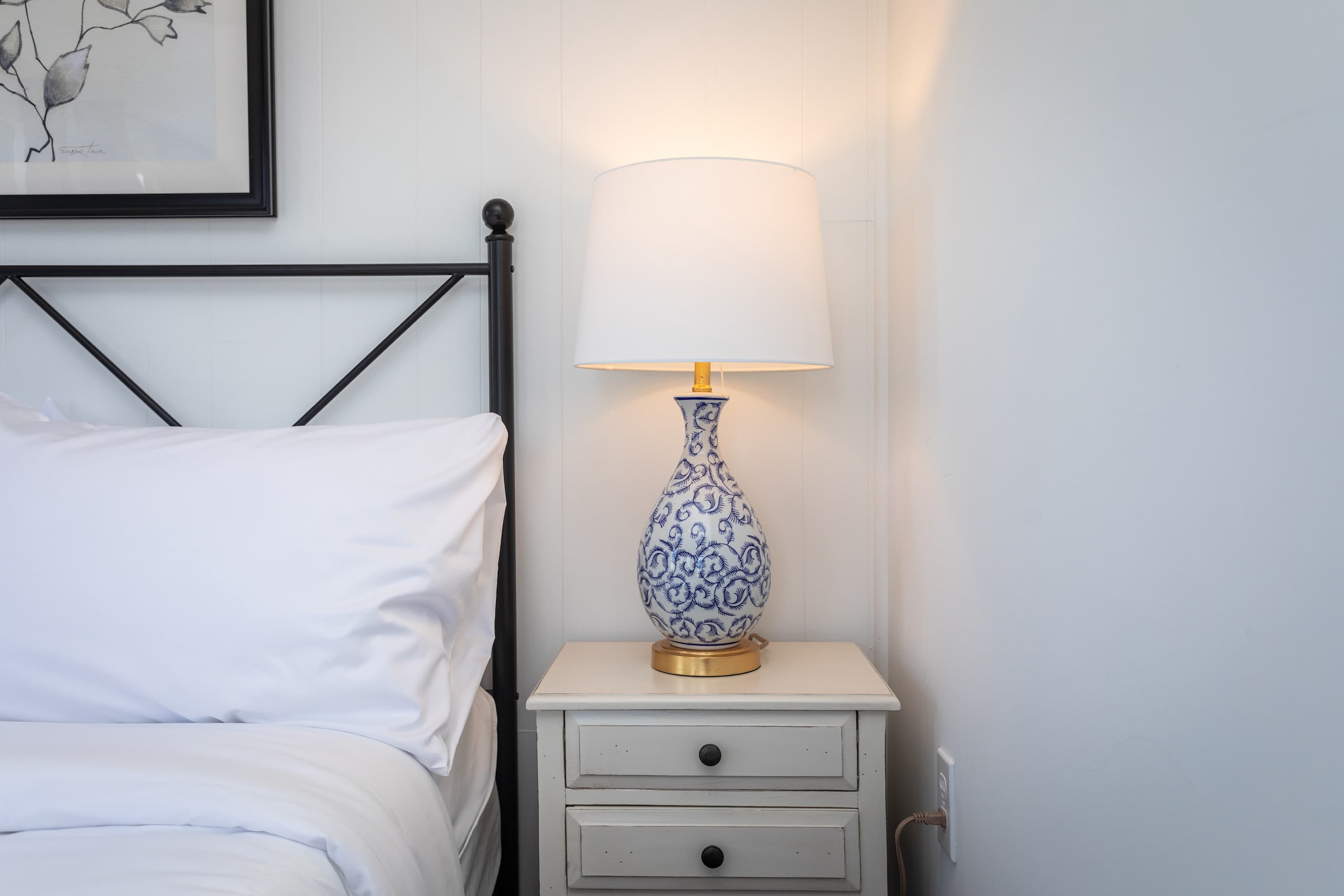 A bedside table with a decorative blue and white lamp next to a bed with white linens.