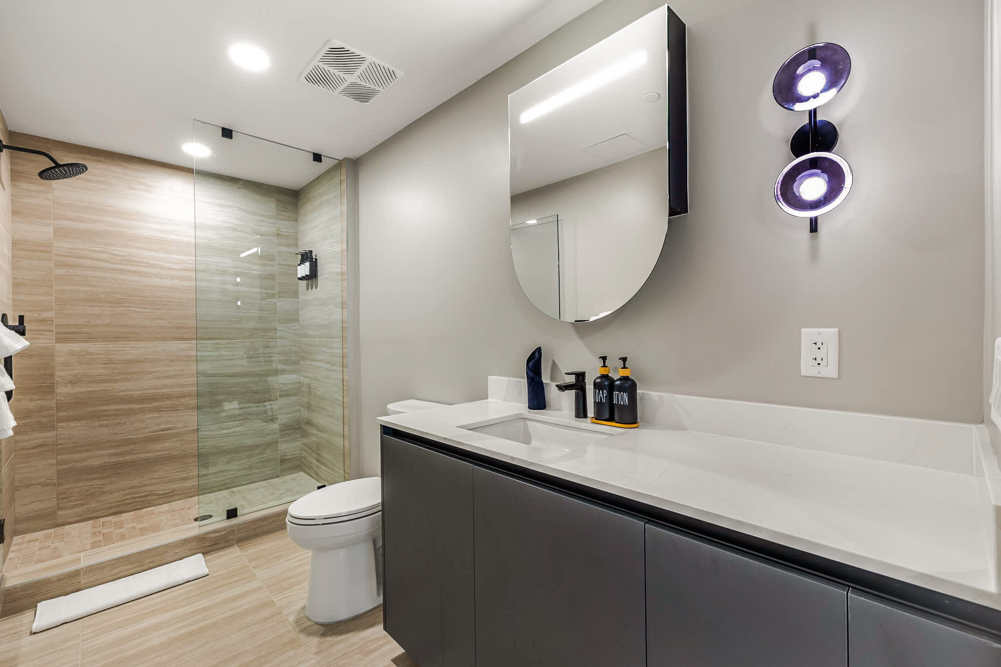 Modern private bathroom with a glass walk-in rain shower, sculptural lighting, rounded mirror, and sleek charcoal vanity.