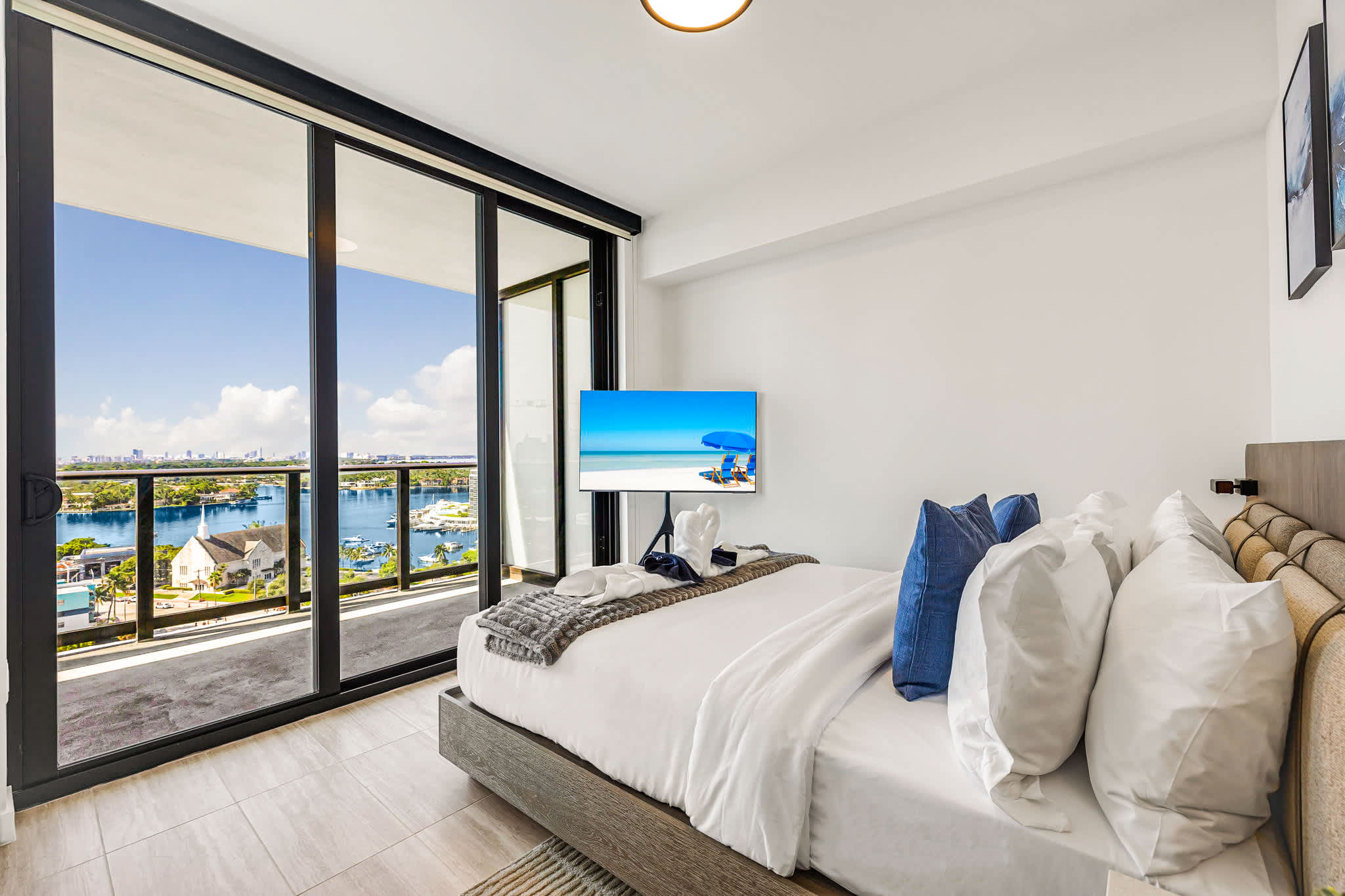 Wake up to sweeping marina and skyline views from a bright bedroom with floor-to-ceiling balcony doors, crisp white bedding, and blue coastal accents.