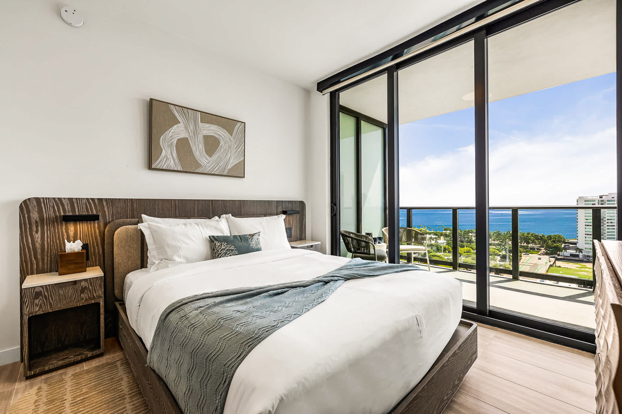 Serene bedroom with crisp linens, a textured blue throw, and floor-to-ceiling glass doors opening to ocean and city views.