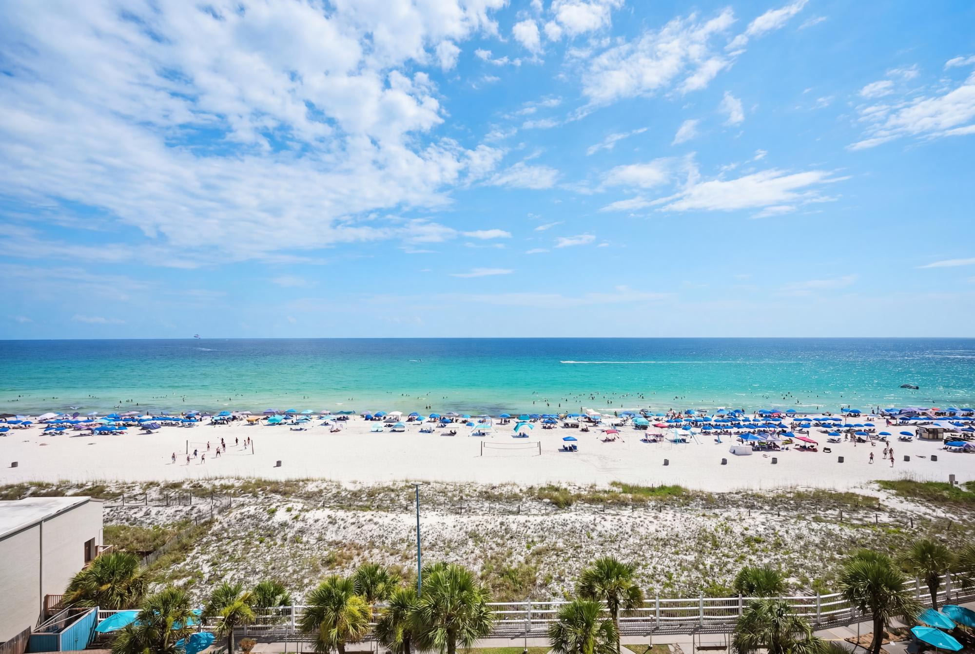 The Summit 830 – View of a sandy beach with umbrellas, turquoise water, and a clear blue sky.