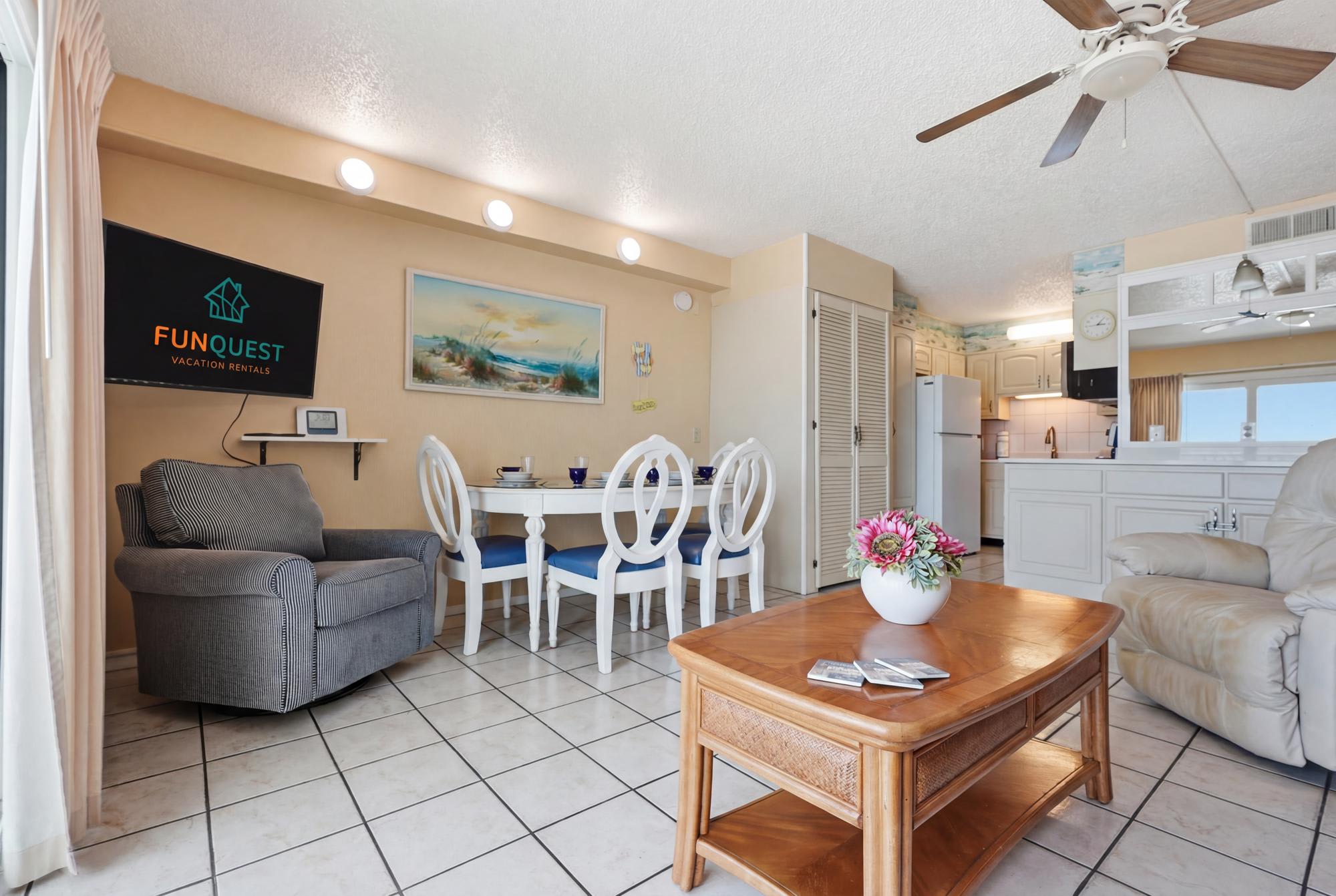 The Summit 830 – Living area with armchair, dining table, kitchenette, and wall-mounted TV. Natural light from window.