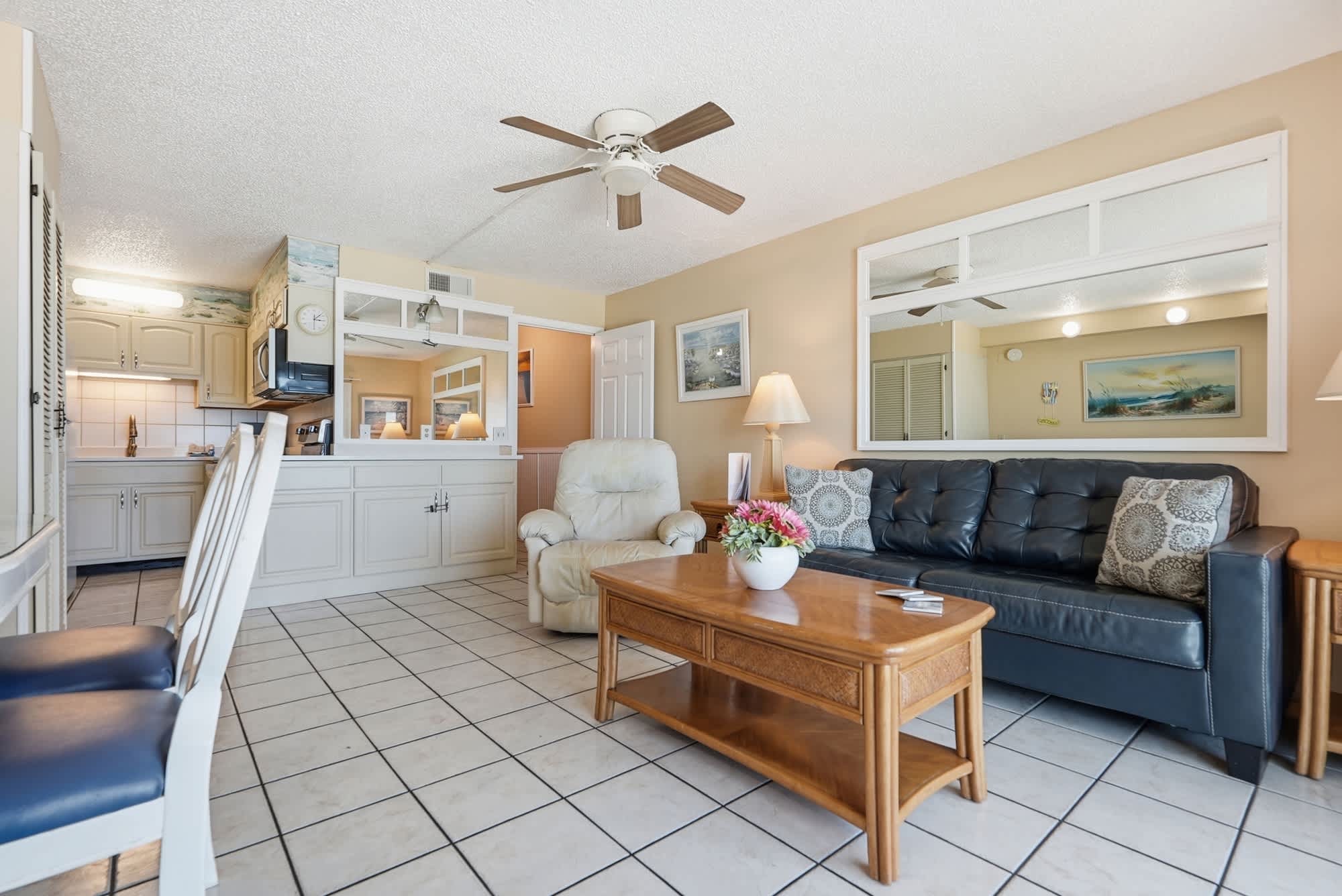 The Summit 830 – Living room with tile floor, black sofa, coffee table, and open kitchen. Light colors and ceiling fan visible.