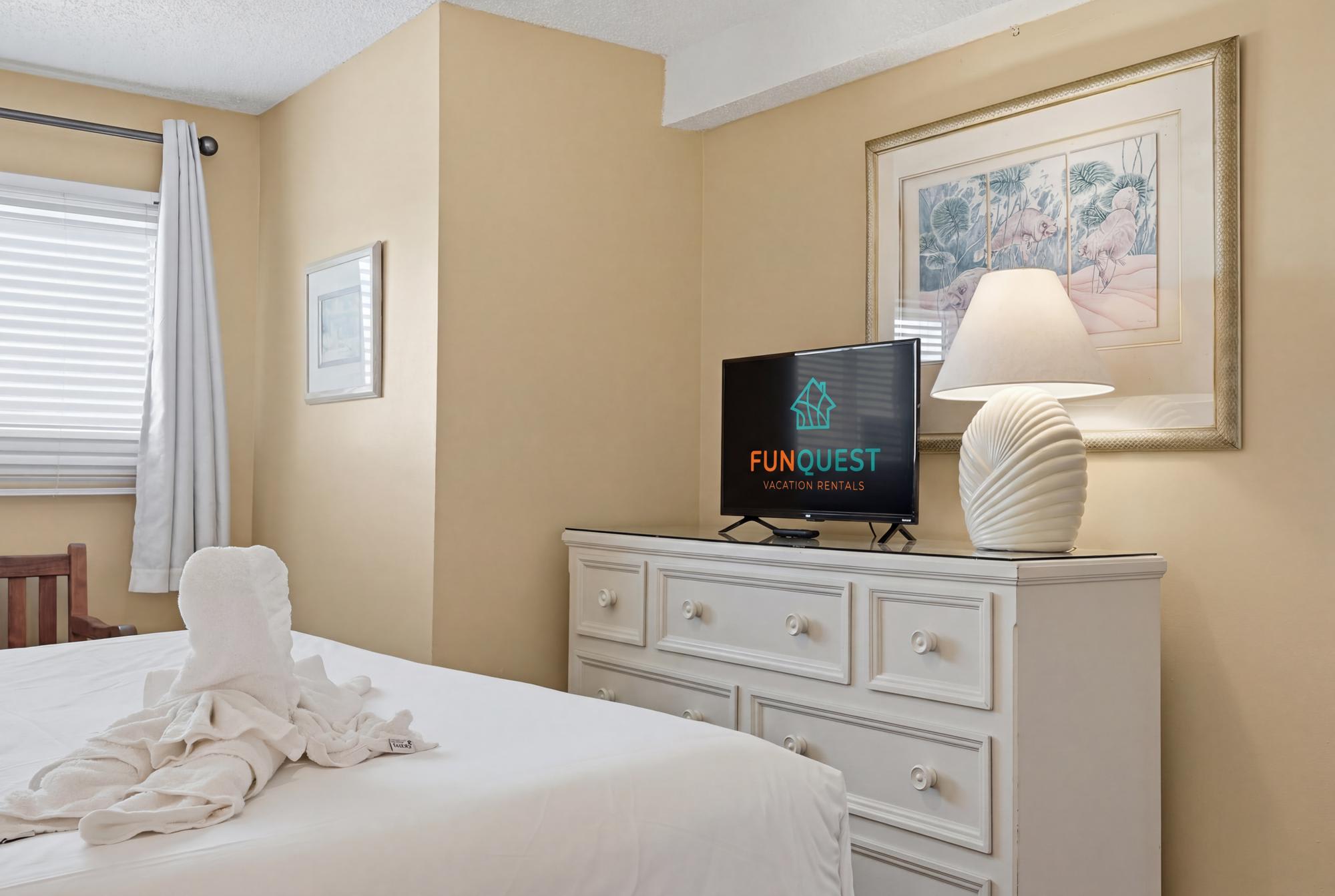 The Summit 830 – Cozy bedroom with a dresser, TV, lamp, and towel art on the bed.