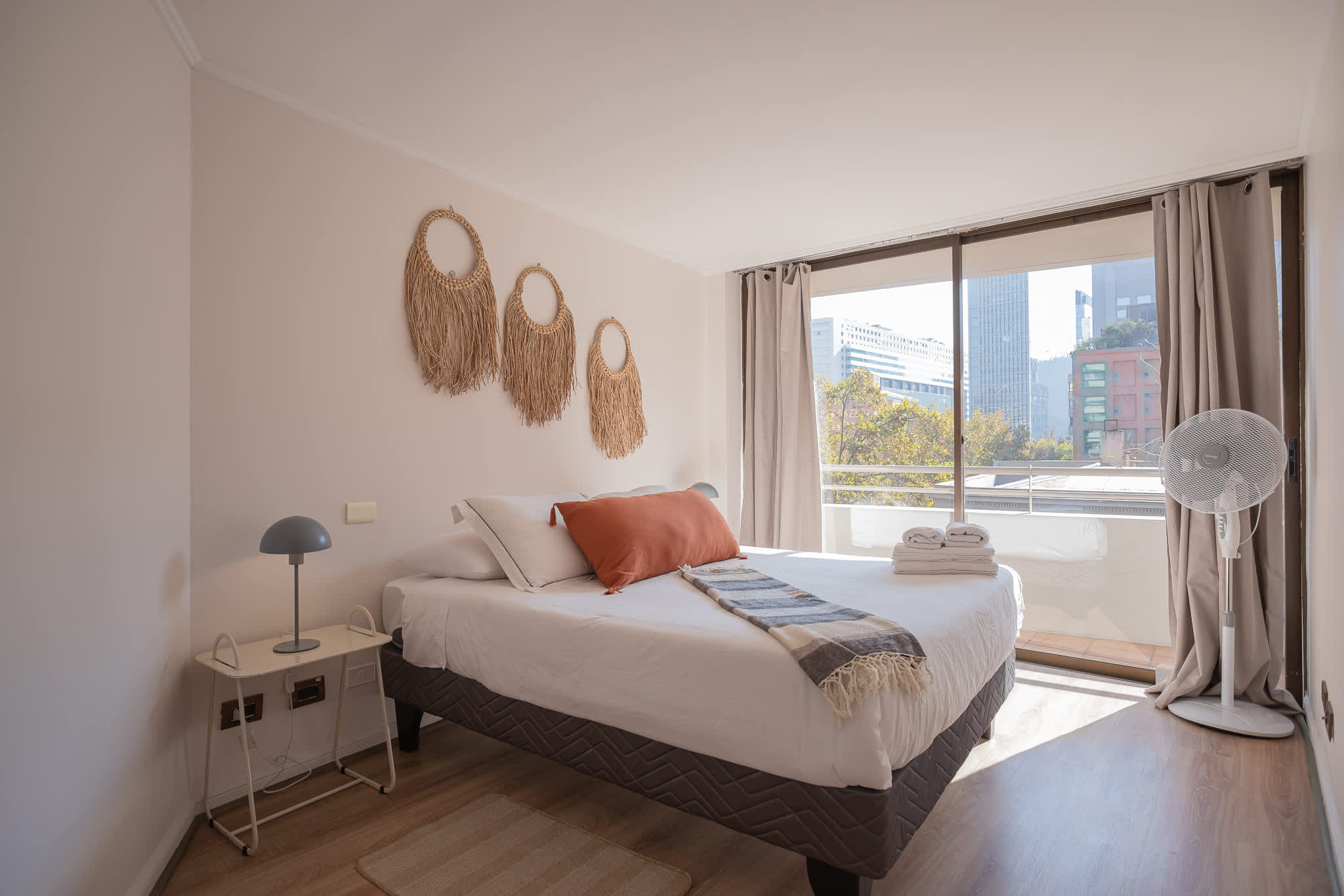 Bright bedroom with a neatly layered bed, woven wall decor, and expansive city views through the sliding glass doors.