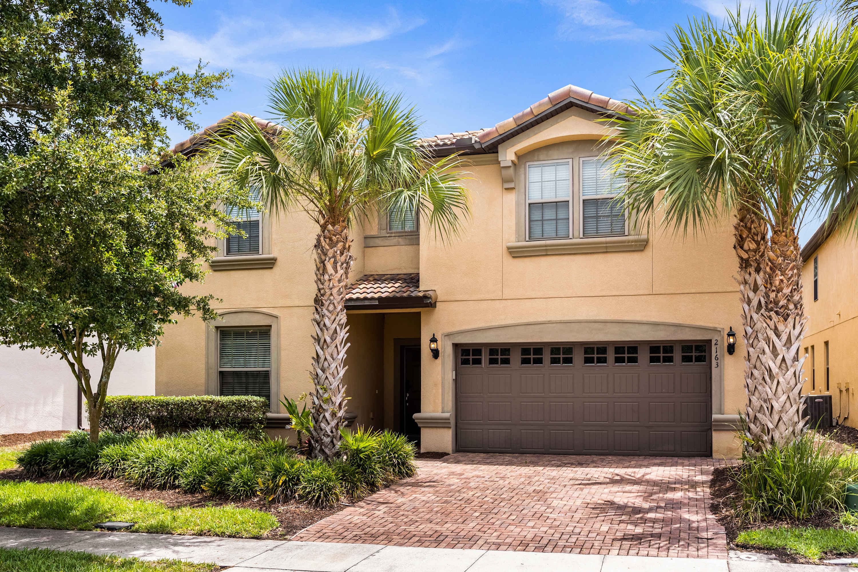 Palm-lined villa entrance with a spacious brick-paver driveway and large two-car garage, welcoming groups with polished landscaping and a sunny Florida-style exterior.