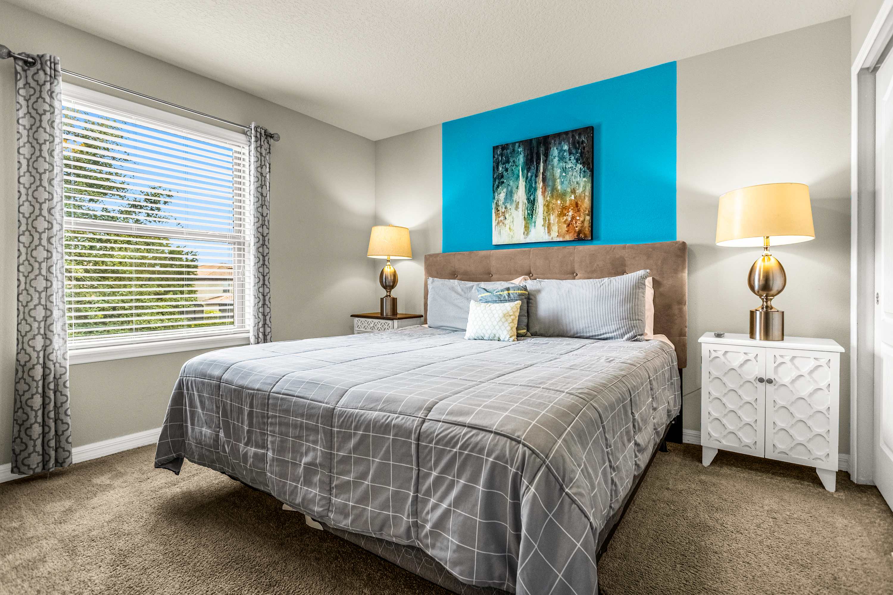 Private bedroom with a plush upholstered bed, vibrant turquoise accent wall, colorful abstract artwork, and a sunny window overlooking leafy trees.
