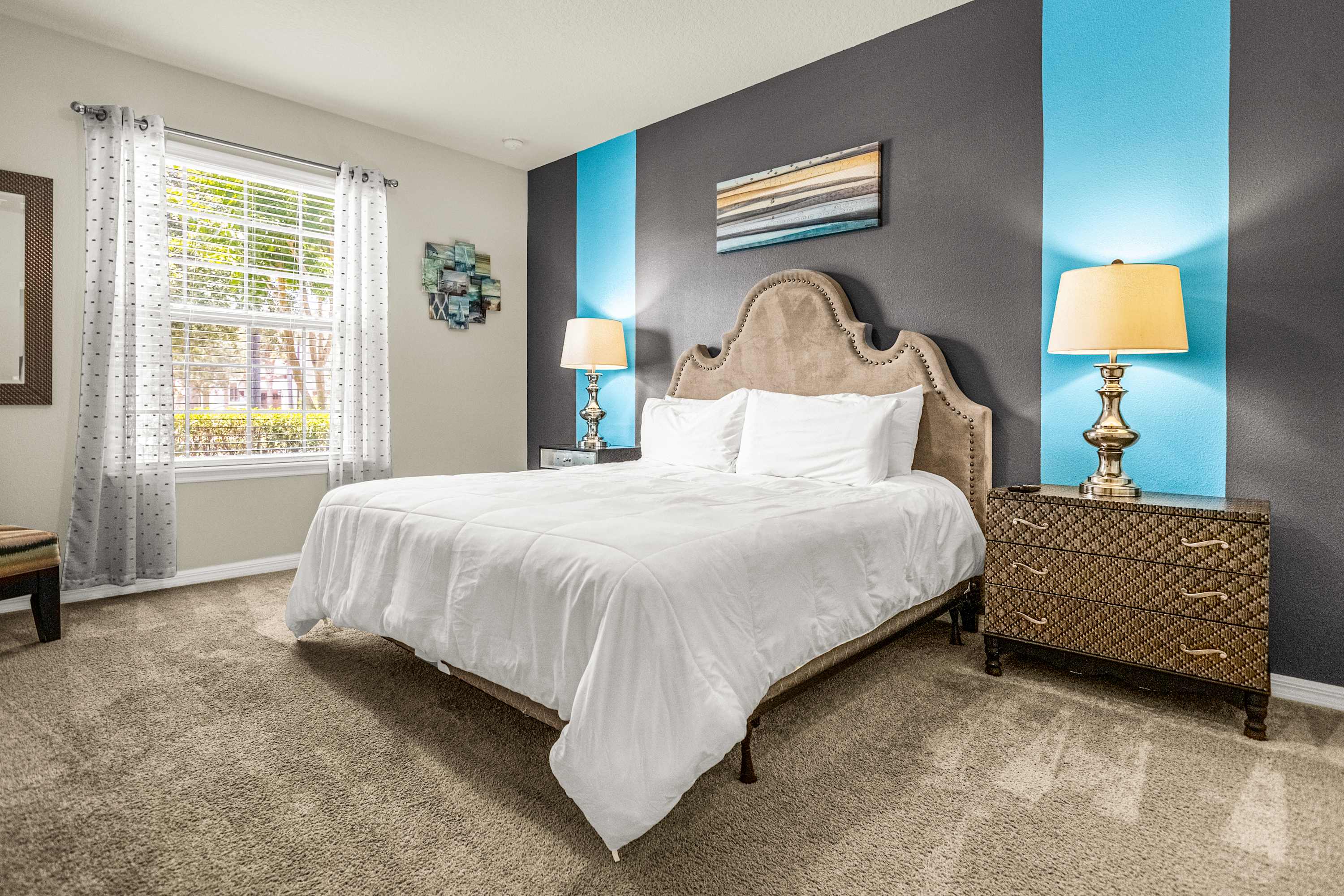Private bedroom with a nailhead-trimmed statement headboard, crisp white bedding, turquoise accent panels, and a sunlit window overlooking leafy greenery.