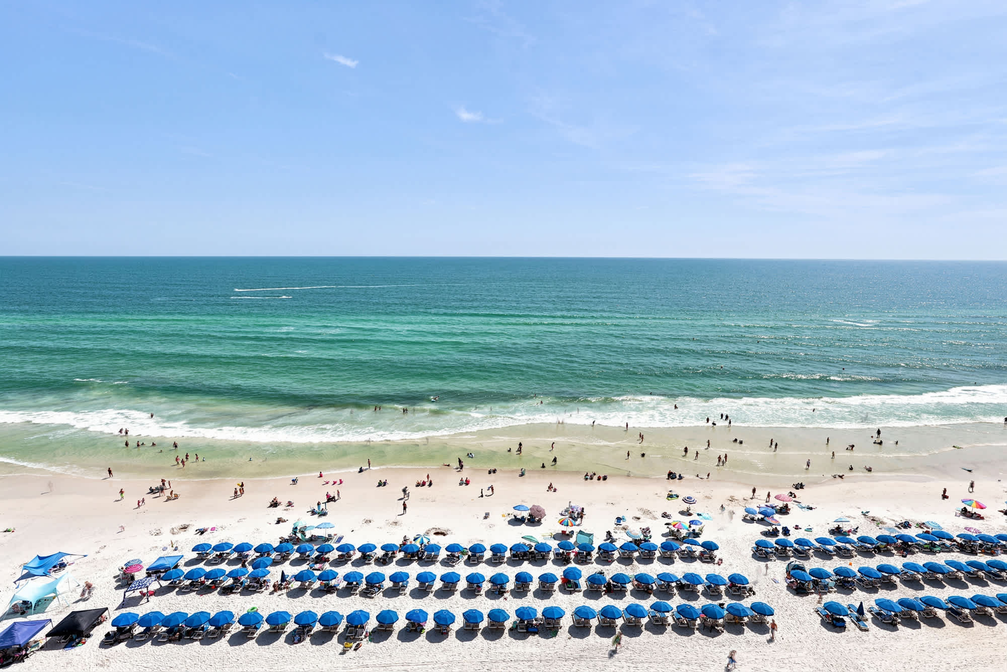 Splash 804E – View of a sandy beach with rows of blue umbrellas and people enjoying the water.