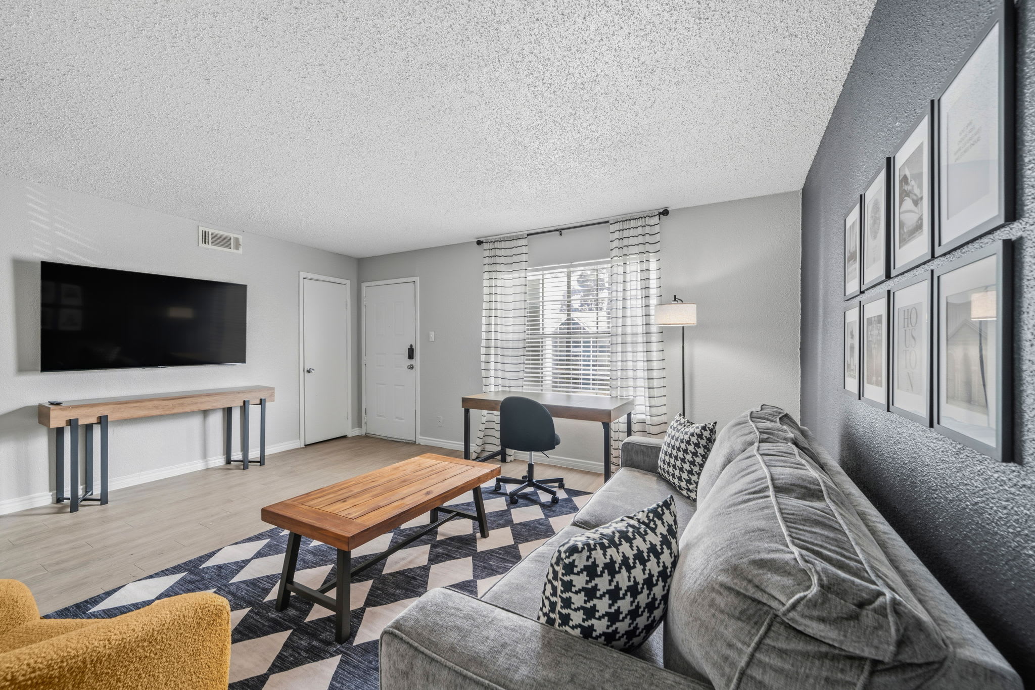 Bright private living room with a large smart TV, comfortable sectional sofa, geometric rug, and a dedicated window-side workspace.
