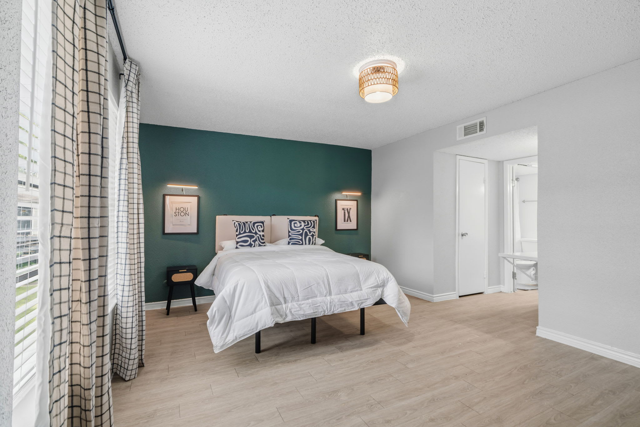 Bright bedroom with a crisp white bed, deep teal accent wall, patterned pillows, and floor-to-ceiling windows framed by plaid curtains.
