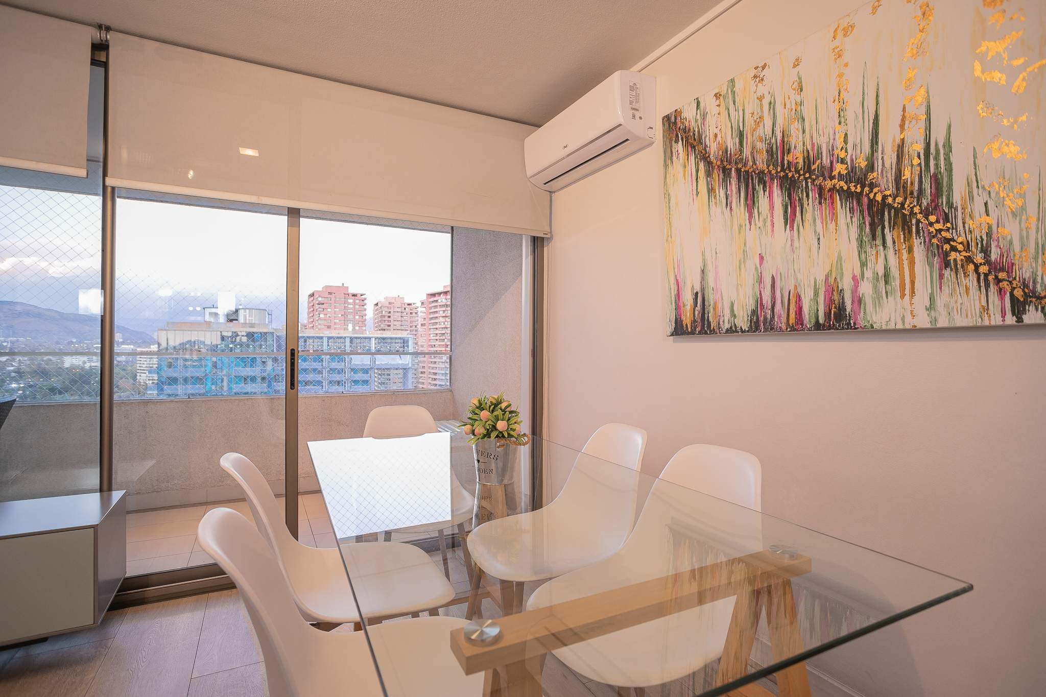Enjoy meals with a stunning city view in this modern dining area, featuring a sleek glass table and vibrant artwork.