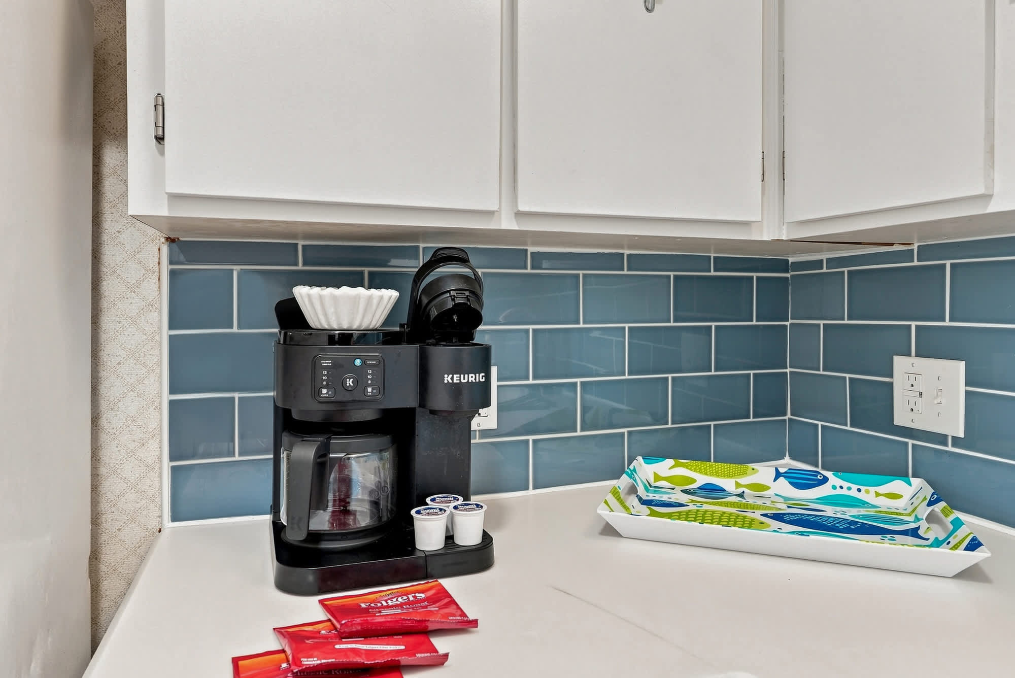 The Summit 308  – Keurig coffee maker with coffee pods on a countertop, blue tile backsplash, and decorative serving tray.