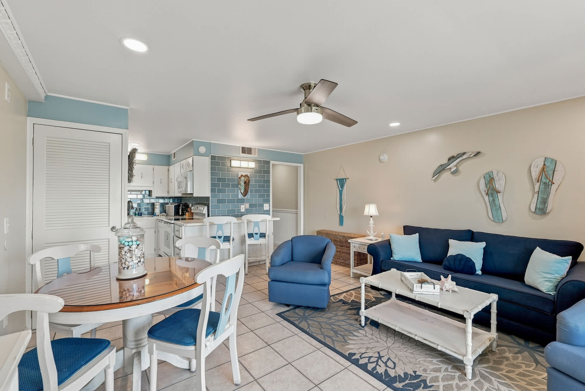The Summit 308  – Living room with blue sofa, dining table, kitchen area, light-colored walls, and beach-themed decor.