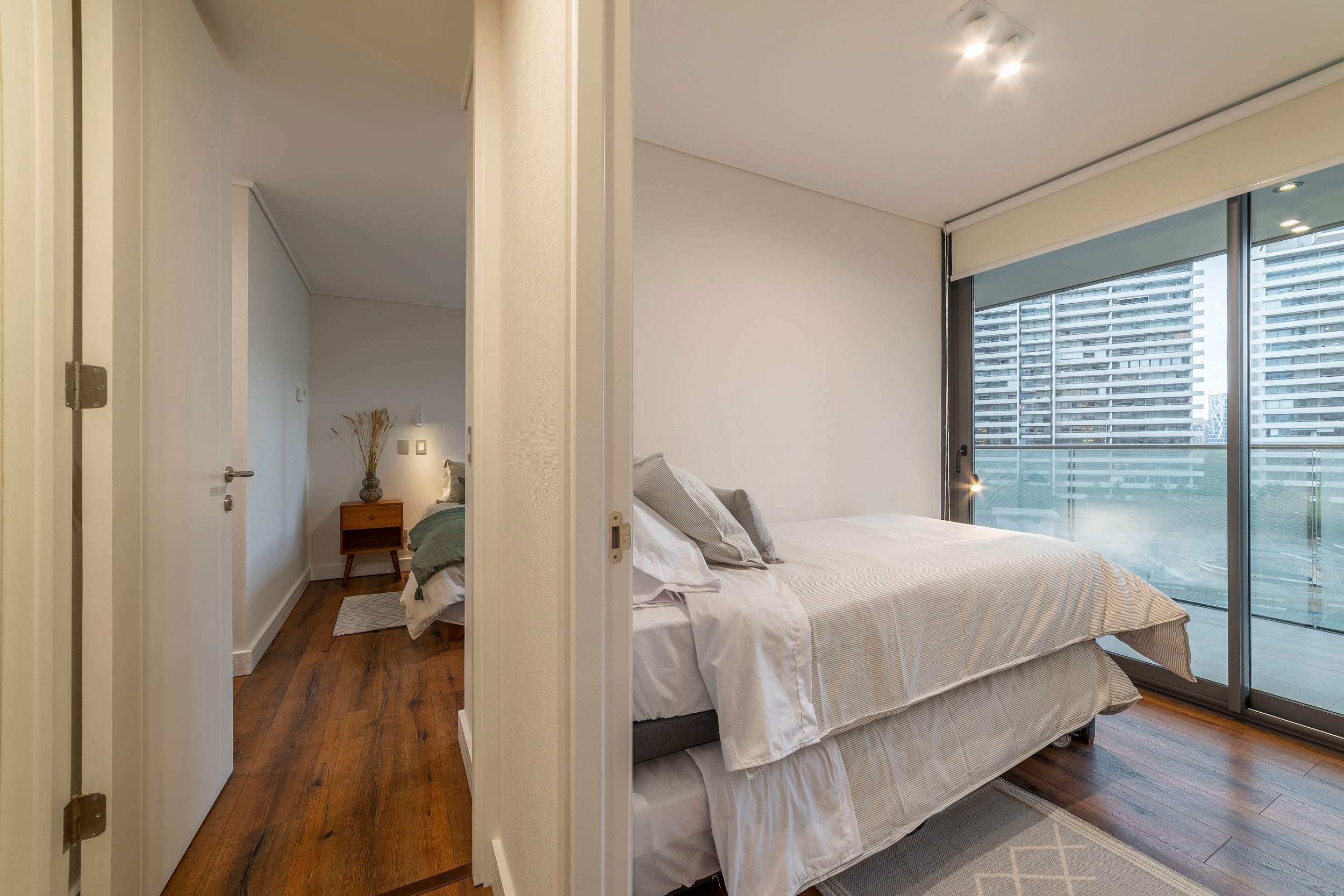 Private bedroom with crisp layered linens, warm wood floors, and floor-to-ceiling windows overlooking Santiago's modern cityscape.