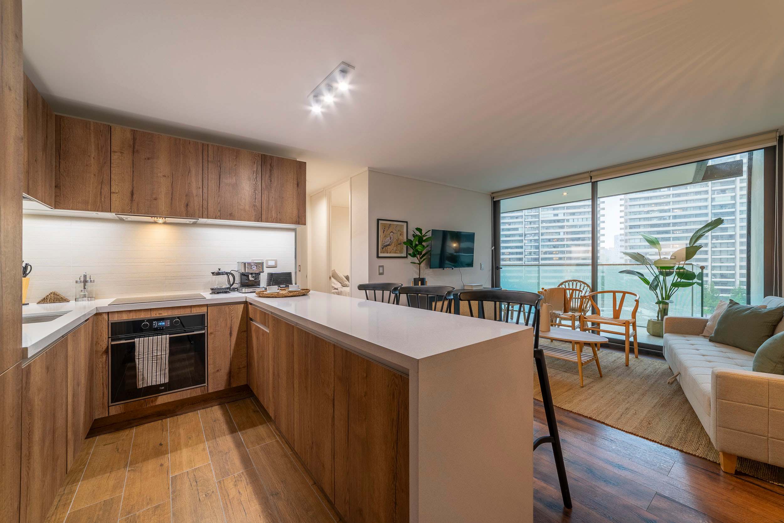 Open-plan kitchen with warm wood cabinetry, a breakfast peninsula, and floor-to-ceiling windows overlooking the city.