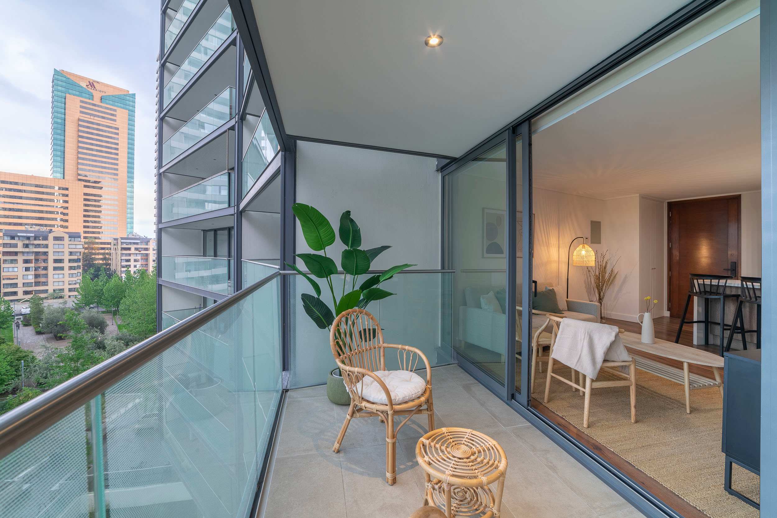 Private balcony with rattan seating, lush greenery, and city views beside a bright open-plan living space.