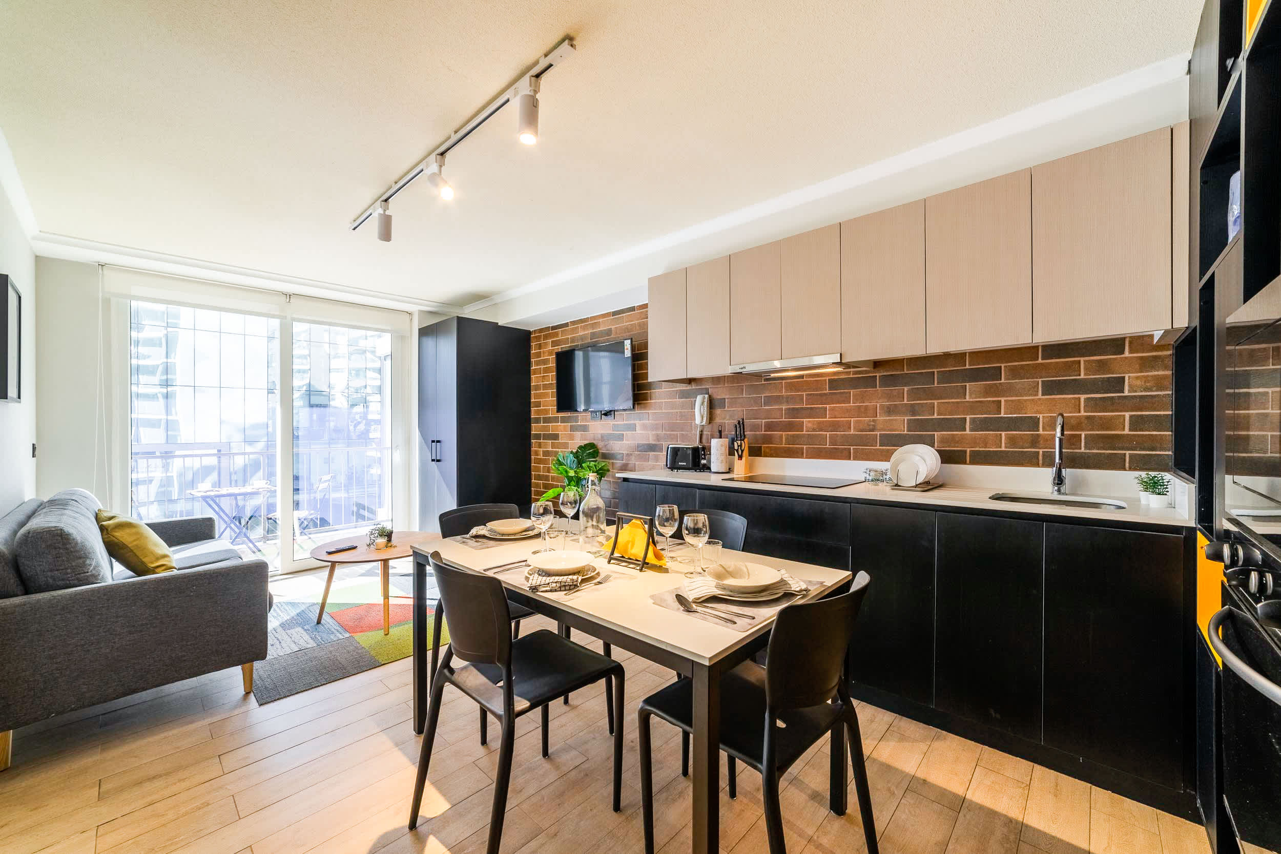 Bright open-plan kitchen and dining area with a warm brick backsplash, modern cabinetry, and a sunlit balcony connection.