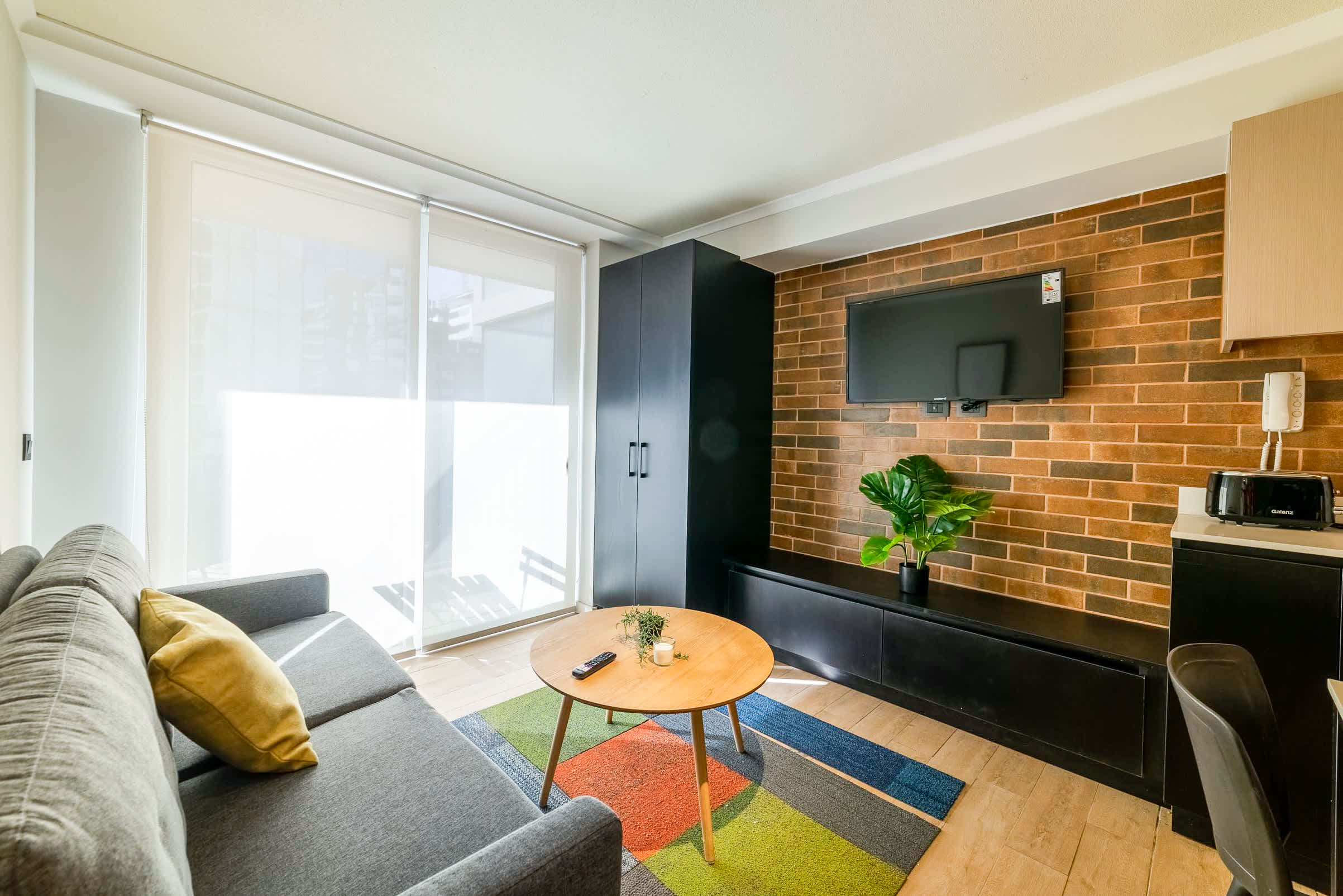 Contemporary studio living room with a gray sofa, colorful geometric rug, wall-mounted TV, and warm exposed-brick accent wall.