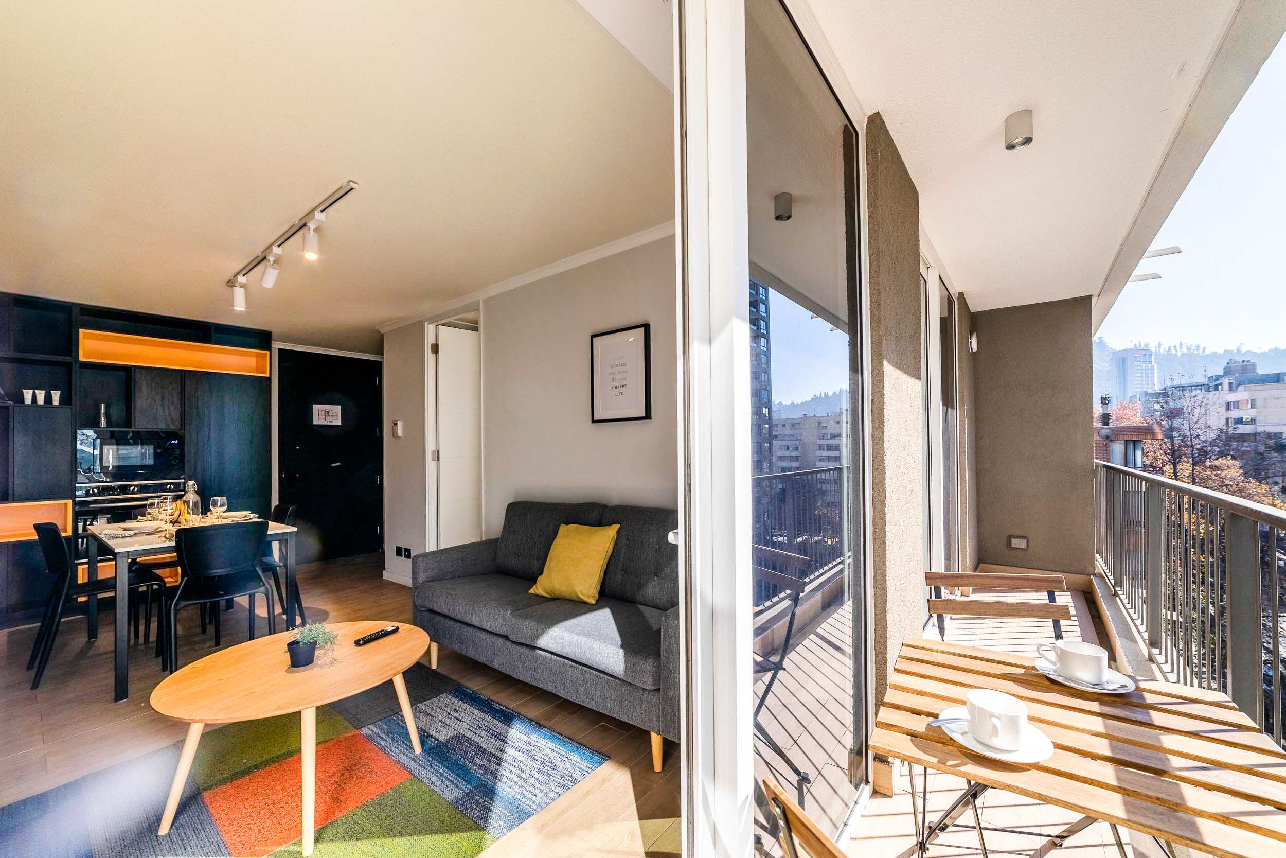 Modern studio living room with a colorful geometric rug, full-height glass doors, and a furnished balcony overlooking the city and surrounding hillside.