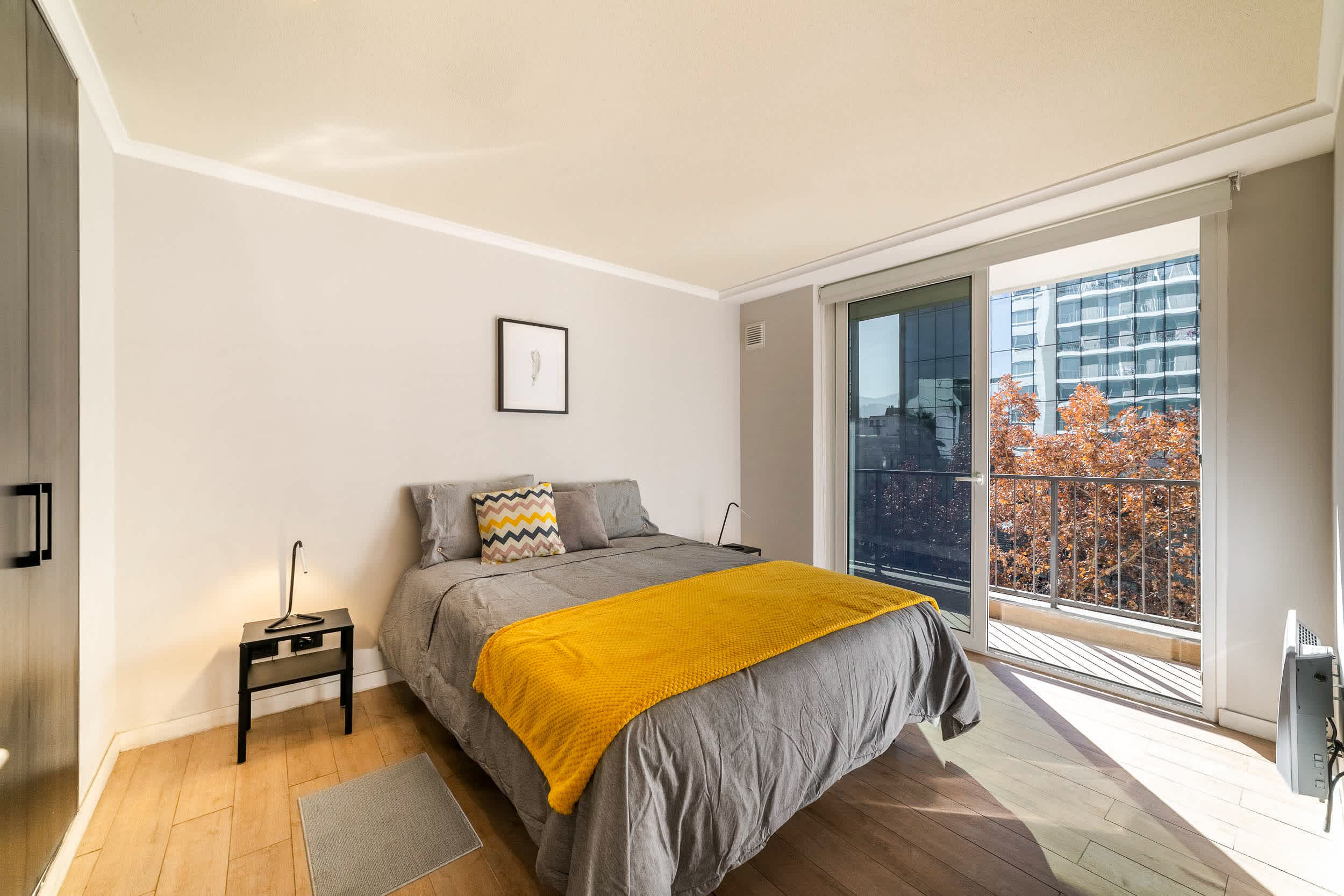 Private bedroom with a mustard-yellow throw and floor-to-ceiling balcony doors overlooking autumn trees and the city.