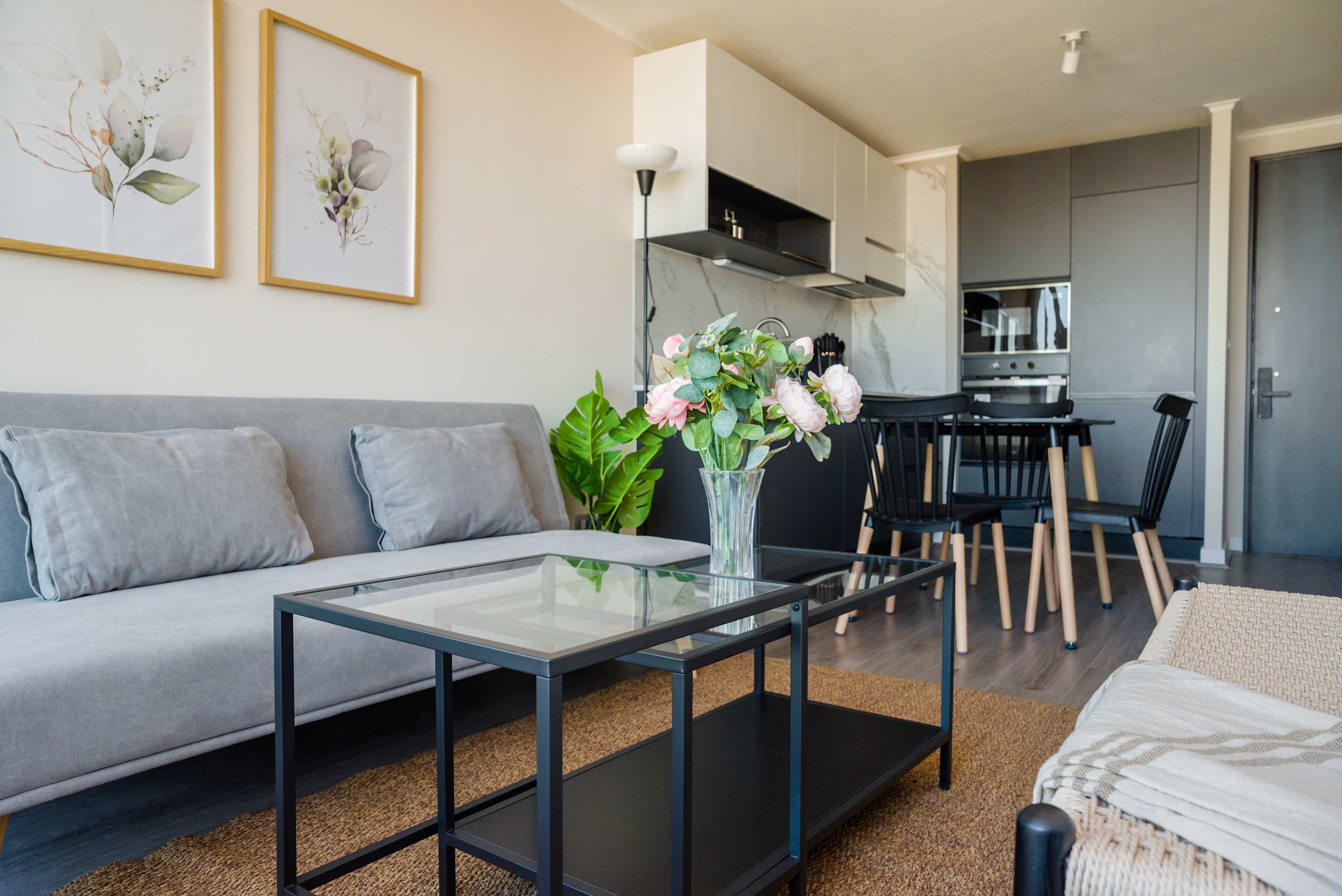 Comfortable living room with a gray sofa, glass coffee tables, and a stylish open kitchen-dining area accented by fresh pink flowers.