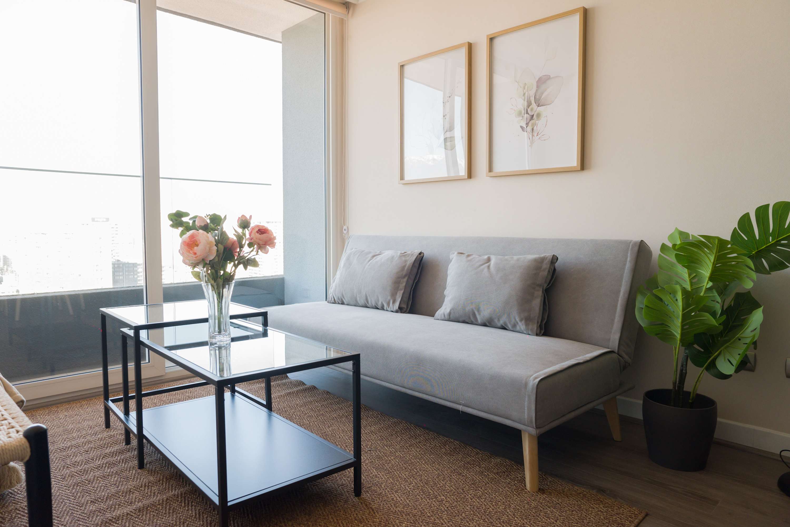 Bright living room with a gray sofa, botanical artwork, and floor-to-ceiling windows overlooking the city.
