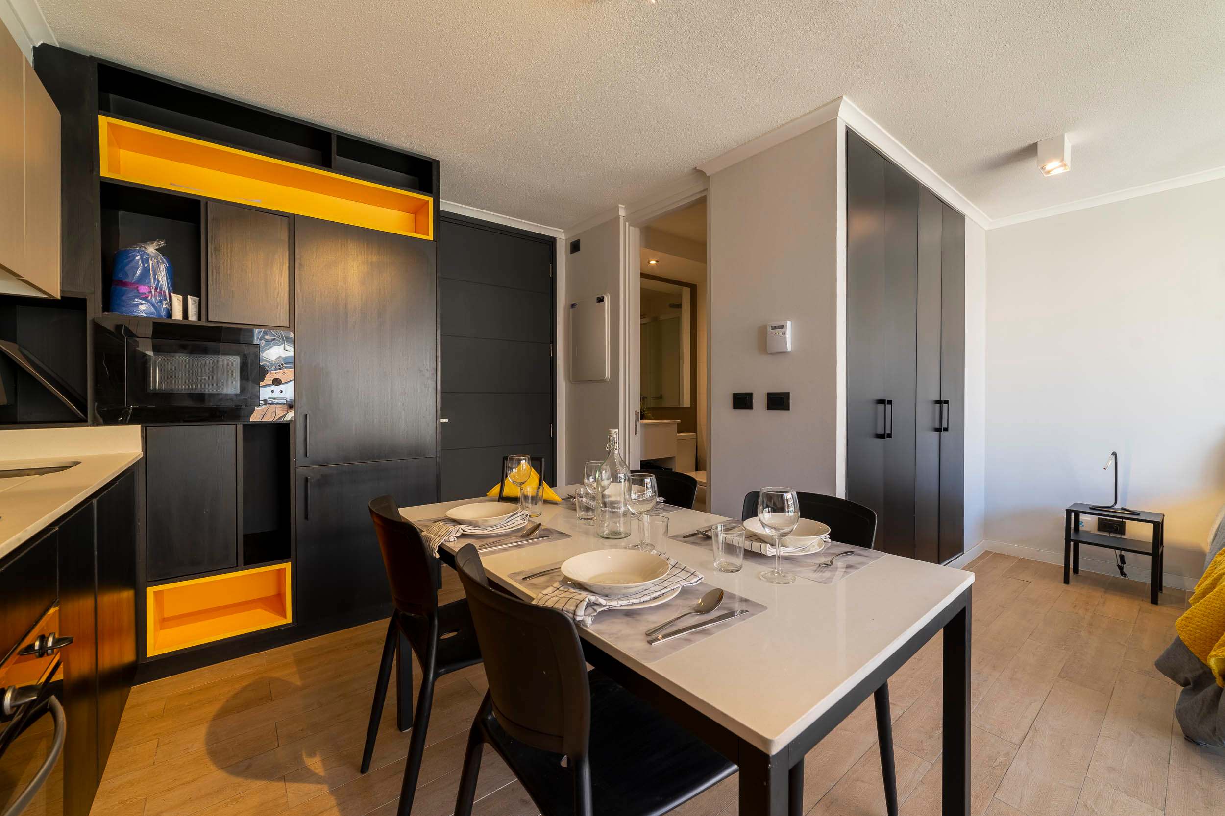 Private dining area with a fully set table, modern black cabinetry, and warm yellow accent shelving beside the compact kitchen.