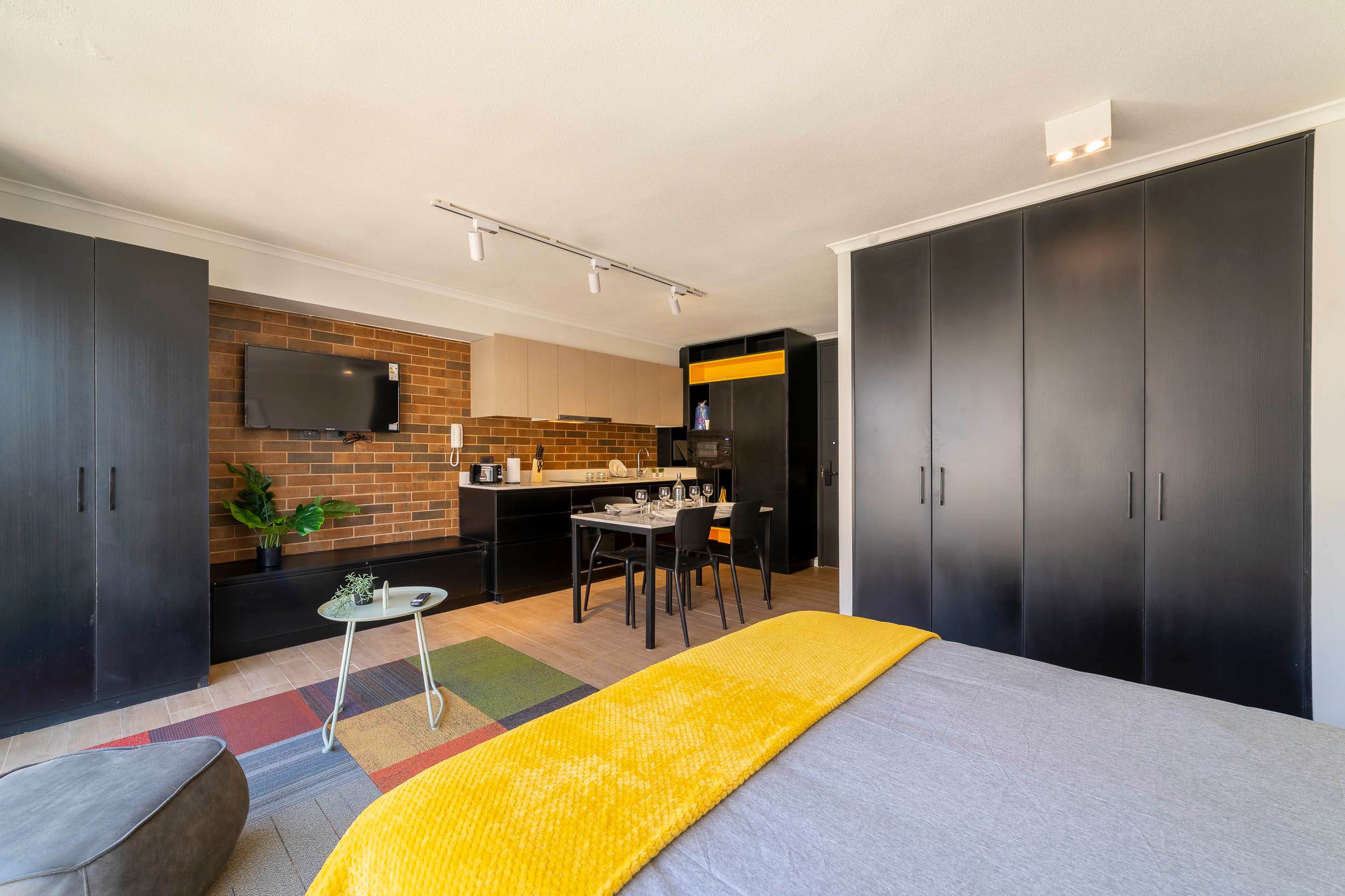 Private studio living space with a mustard throw, exposed brick TV wall, full kitchen, and dining table set for two.