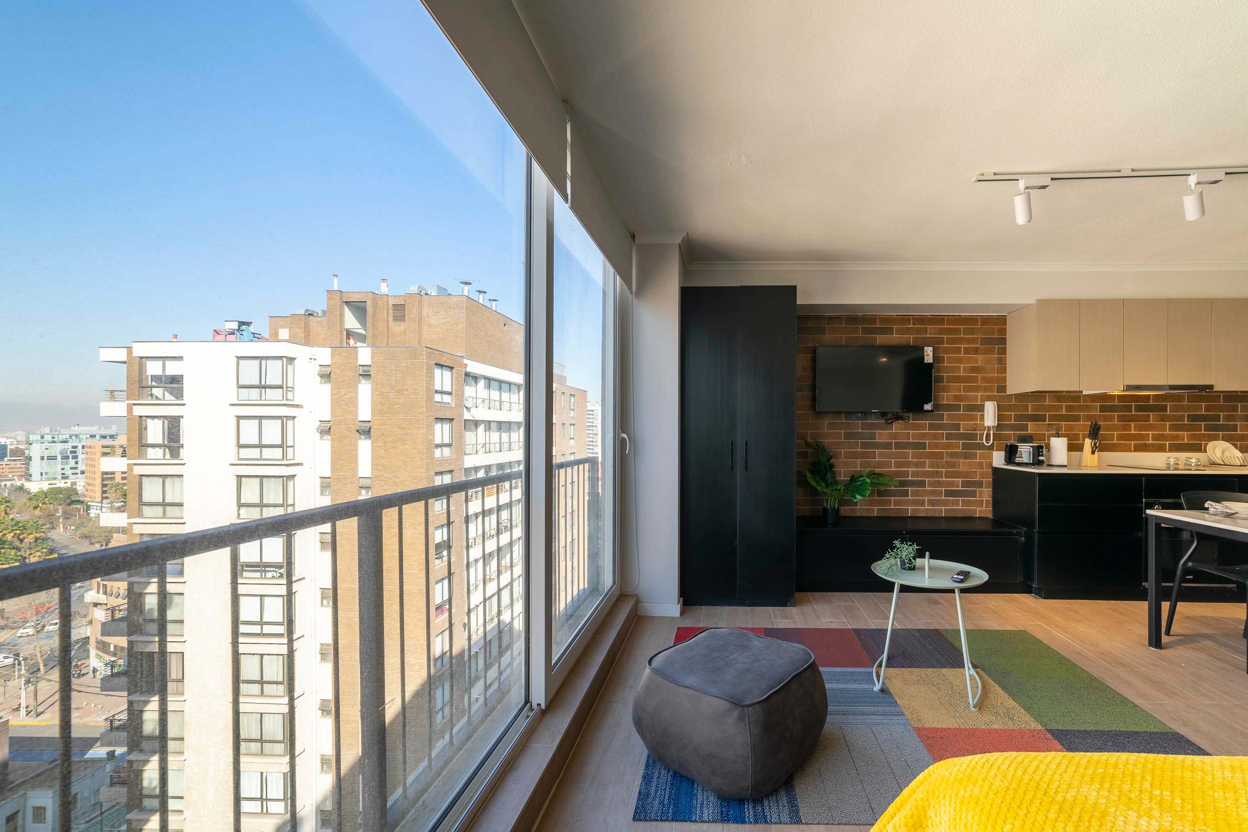 Private studio living room with floor-to-ceiling windows, an urban city view, and a sleek kitchen with a warm brick backsplash.