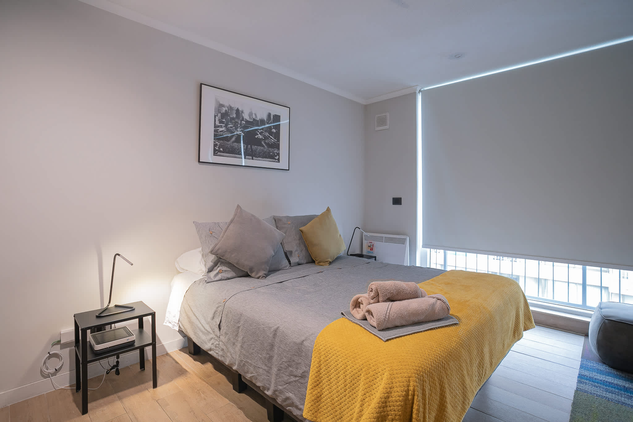 Studio sleeping area with layered gray bedding, mustard throw, rolled towels, and an urban window view.