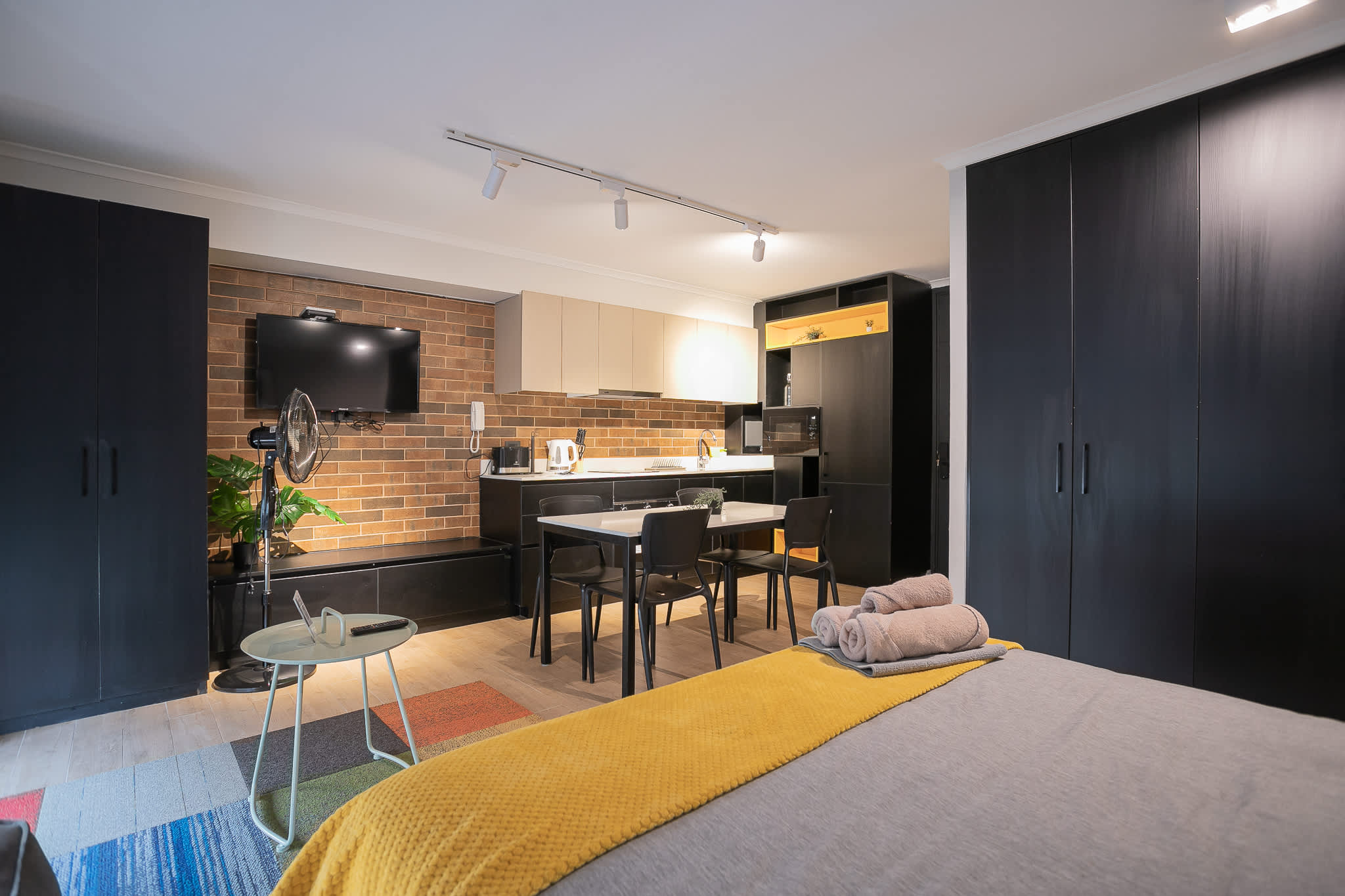 Open-plan studio kitchen with a brick backsplash, full cooking essentials, and a cozy dining area beside the neatly made bed.
