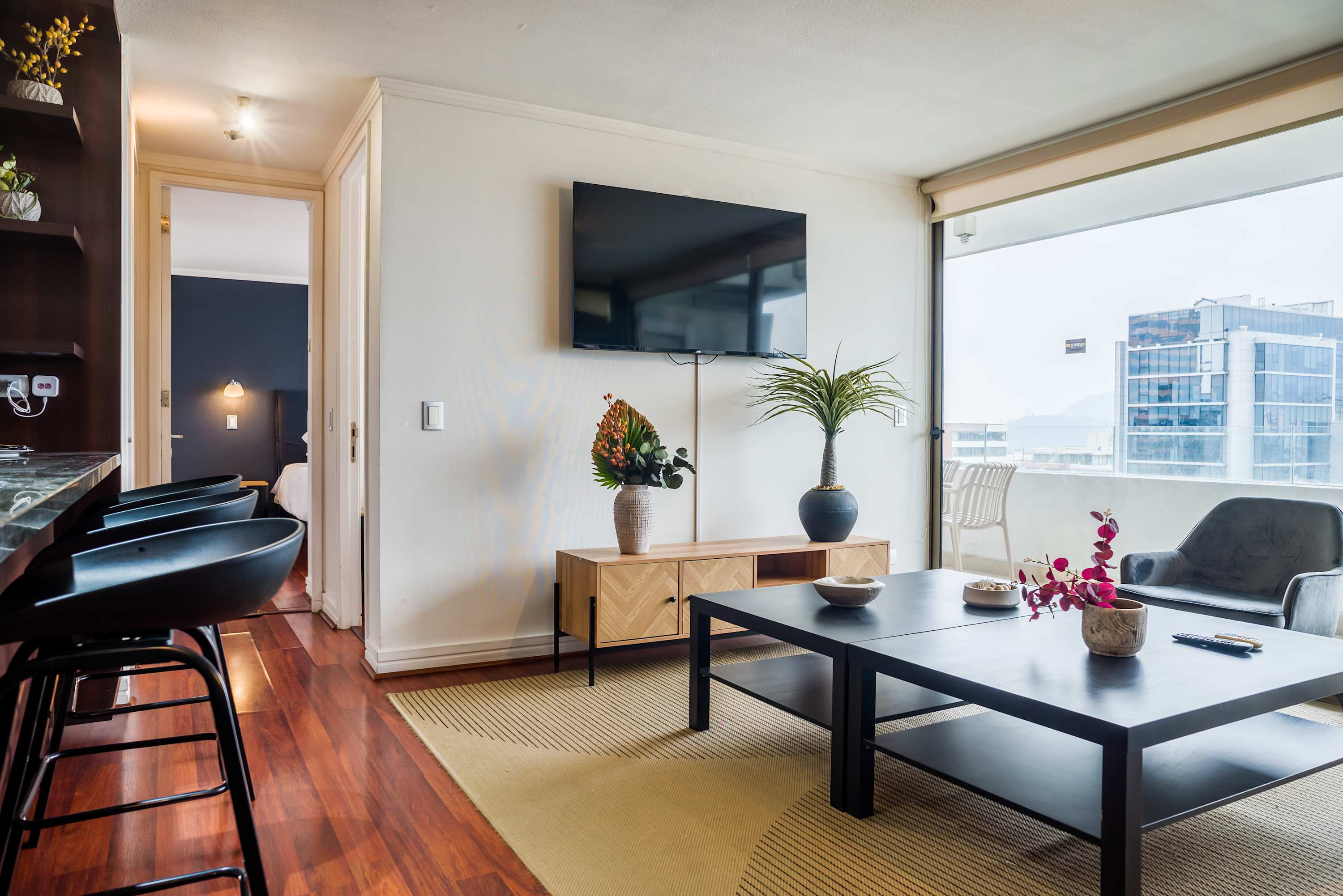 Bright living room with a wall-mounted TV, sculptural decor, and floor-to-ceiling balcony windows overlooking the city.
