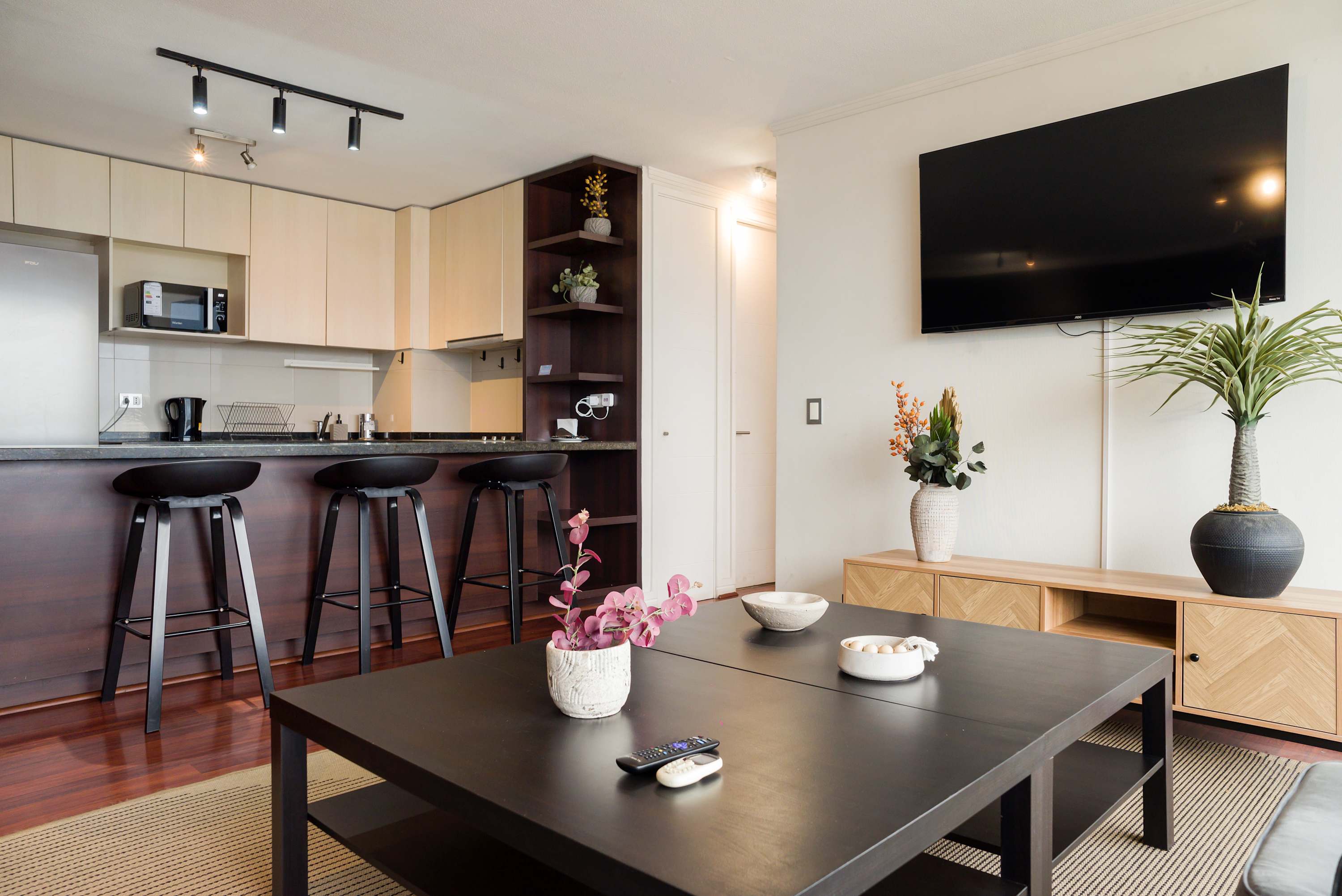 Private open-plan living room with a wall-mounted TV, stylish dark coffee table, and a fully equipped kitchen with three bar stools.