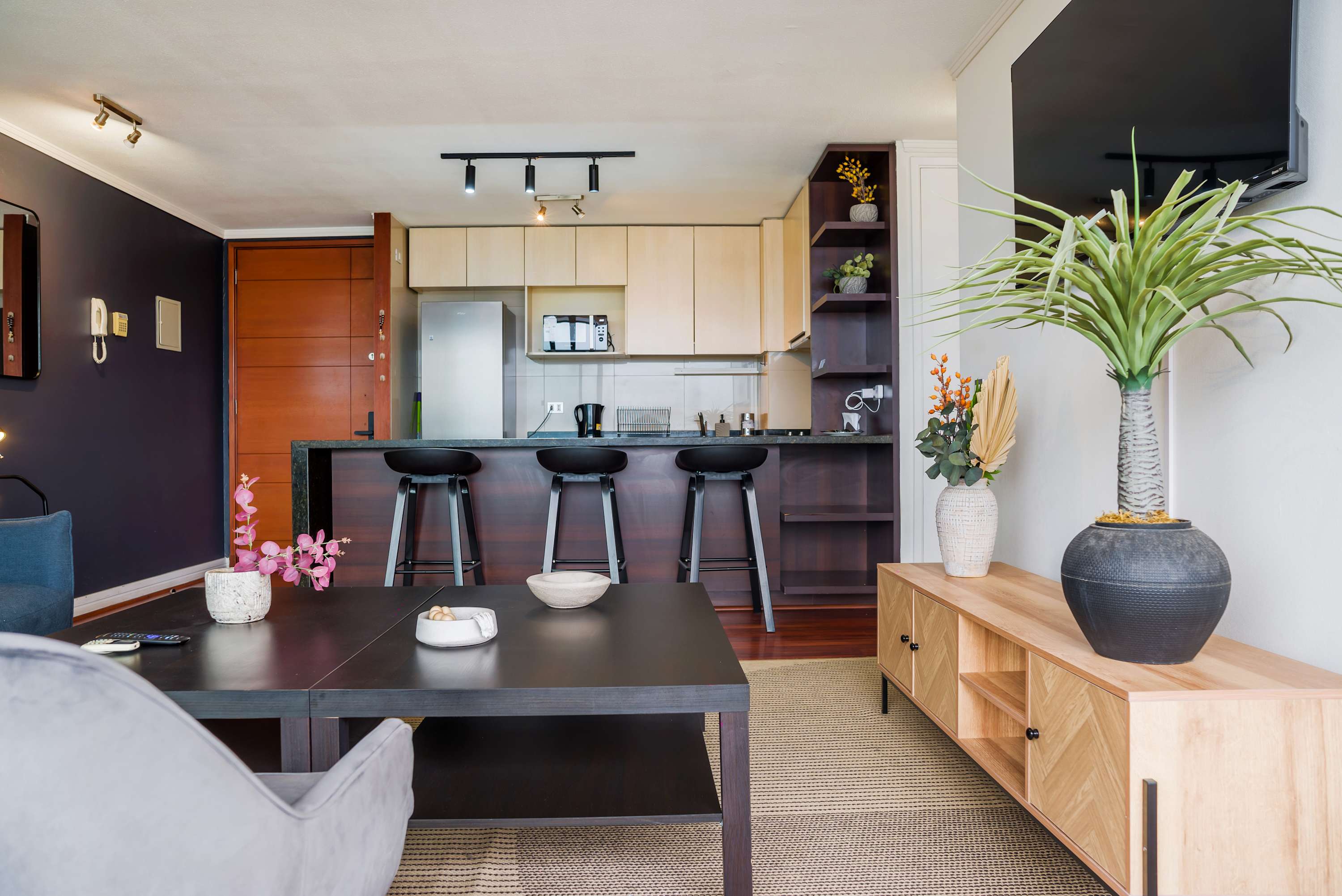 Open-plan kitchen with a dark stone peninsula, three bar stools, and warm wood cabinetry adjoining the living area.