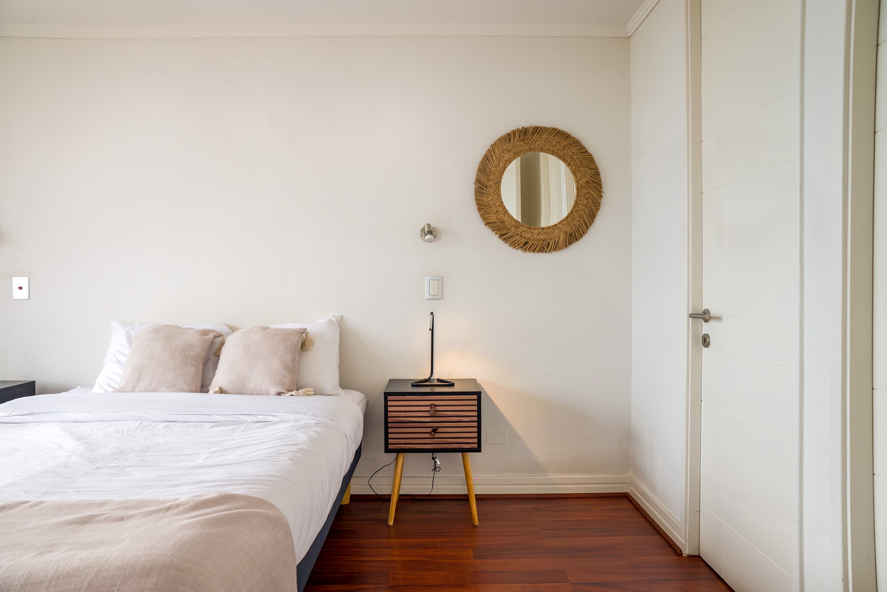 Private bedroom with crisp white bedding, soft beige pillows, a woven round mirror, and warm hardwood floors.
