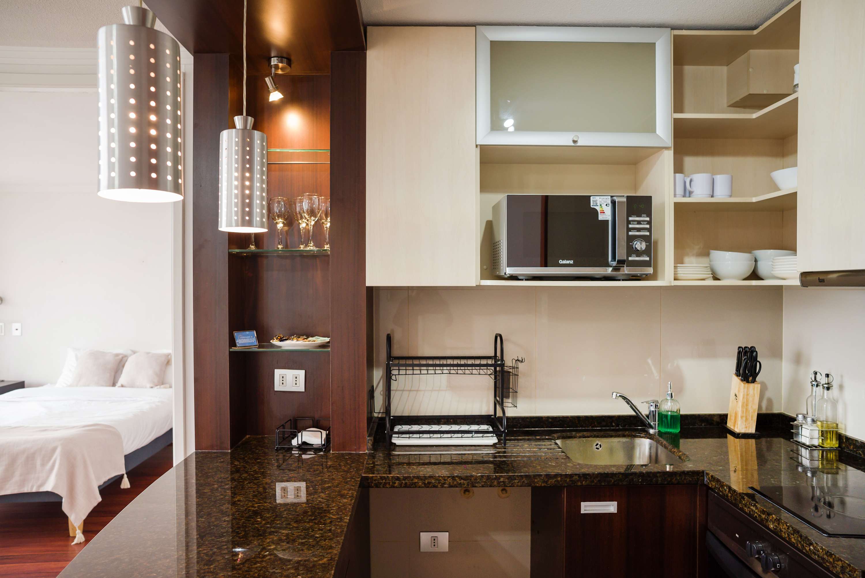 Studio kitchen with granite countertops, open dishware shelving, a microwave, and a glimpse of the neatly made sleeping area.