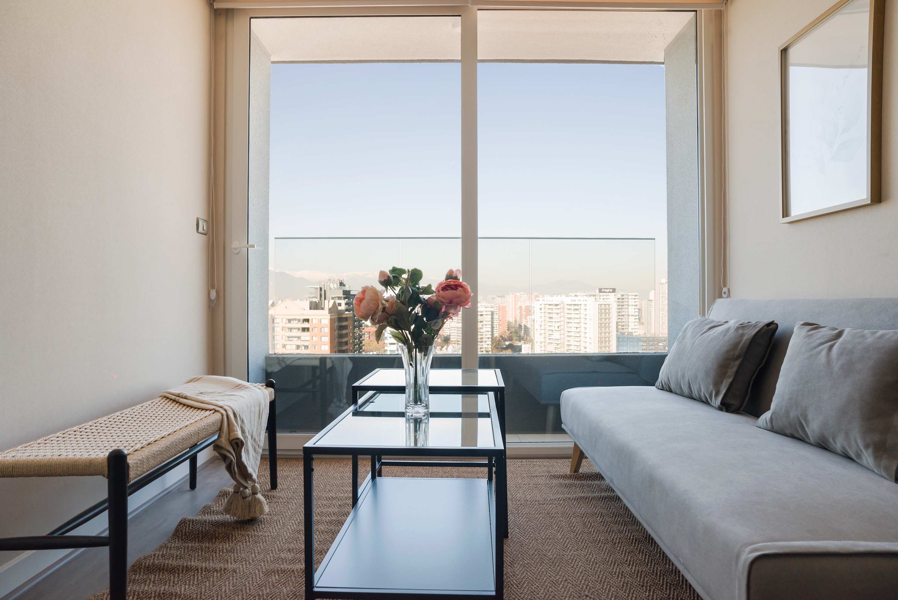 Private living room with a gray sofa, pink floral centerpiece, and sweeping Santiago skyline and mountain views through full-height glass doors.